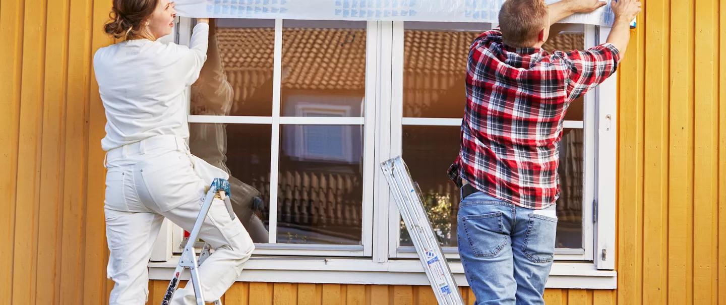 Two people applying protective film above windows on a yellow house.