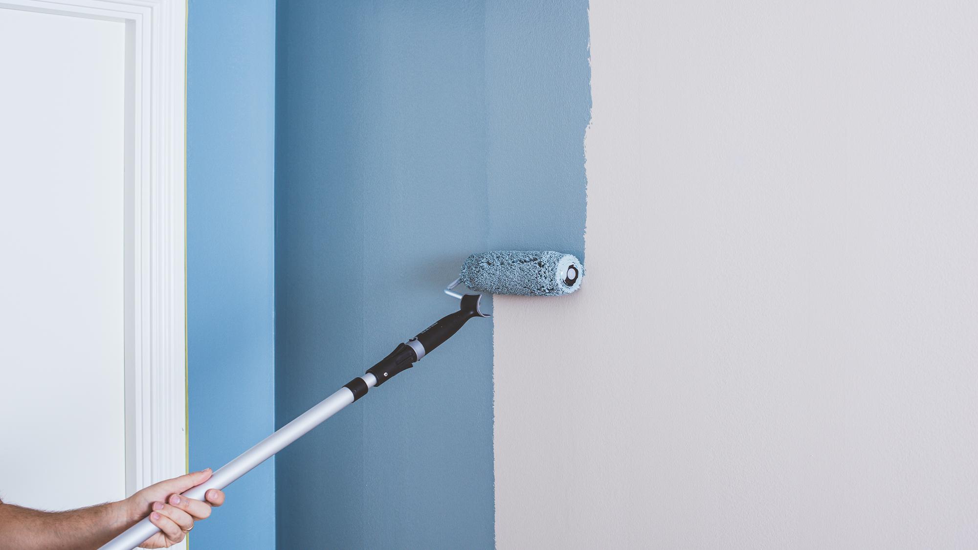 A long-handled paint roller applies blue paint to a light-colored wall.