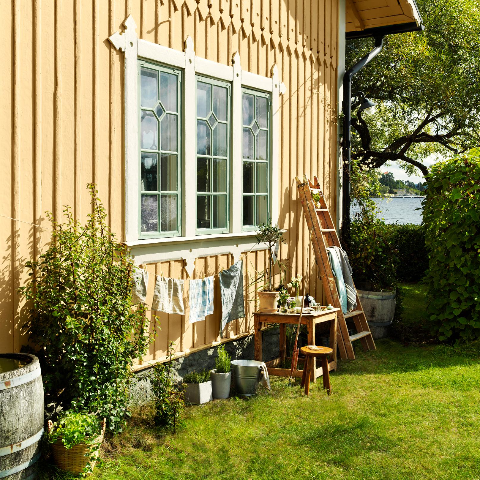 A yellow wooden house with green windows, a garden with an outdoor table, laundry hanging, and a lake view.