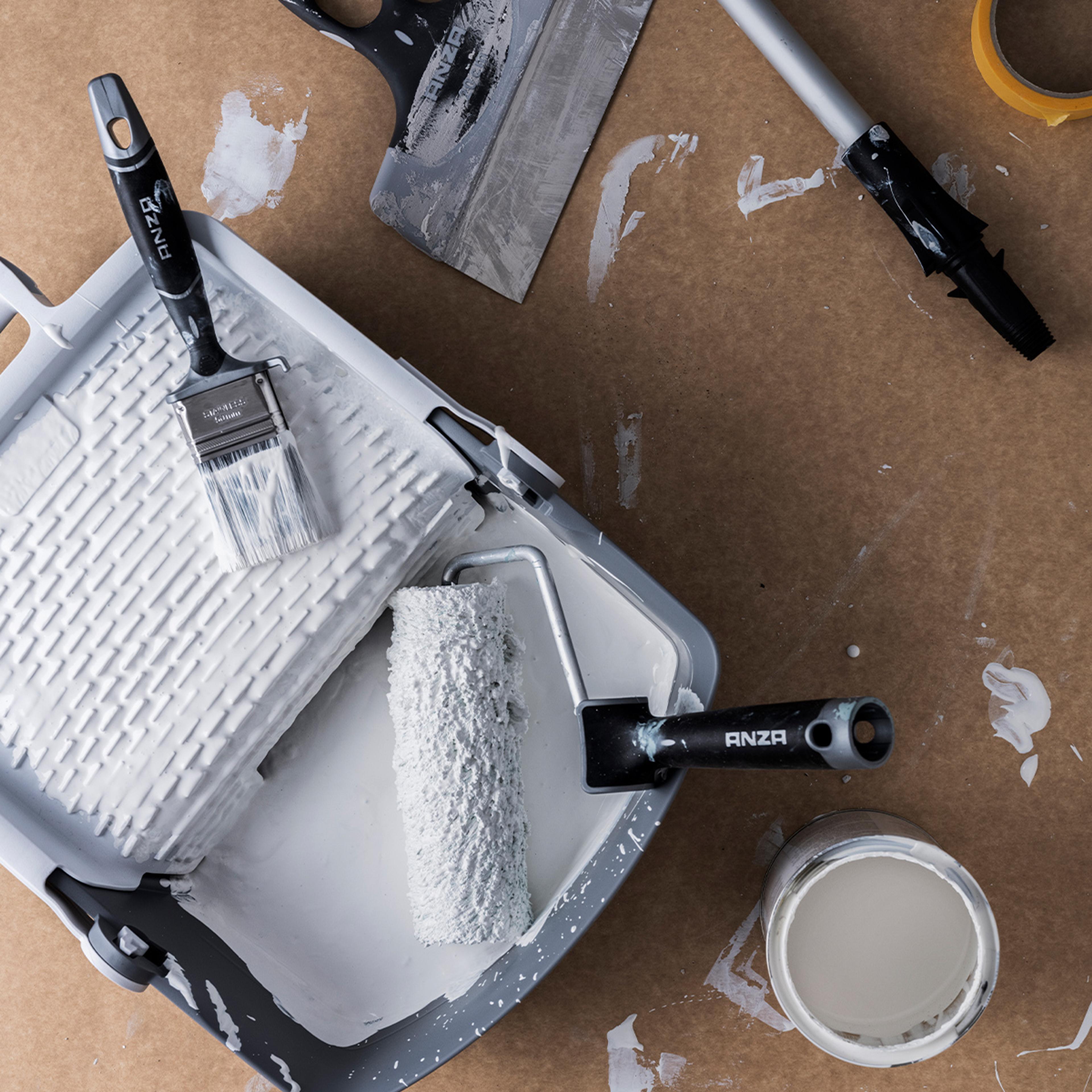 Paint roller and brush in a tray of white paint, with a paint can and splatters on a brown surface.