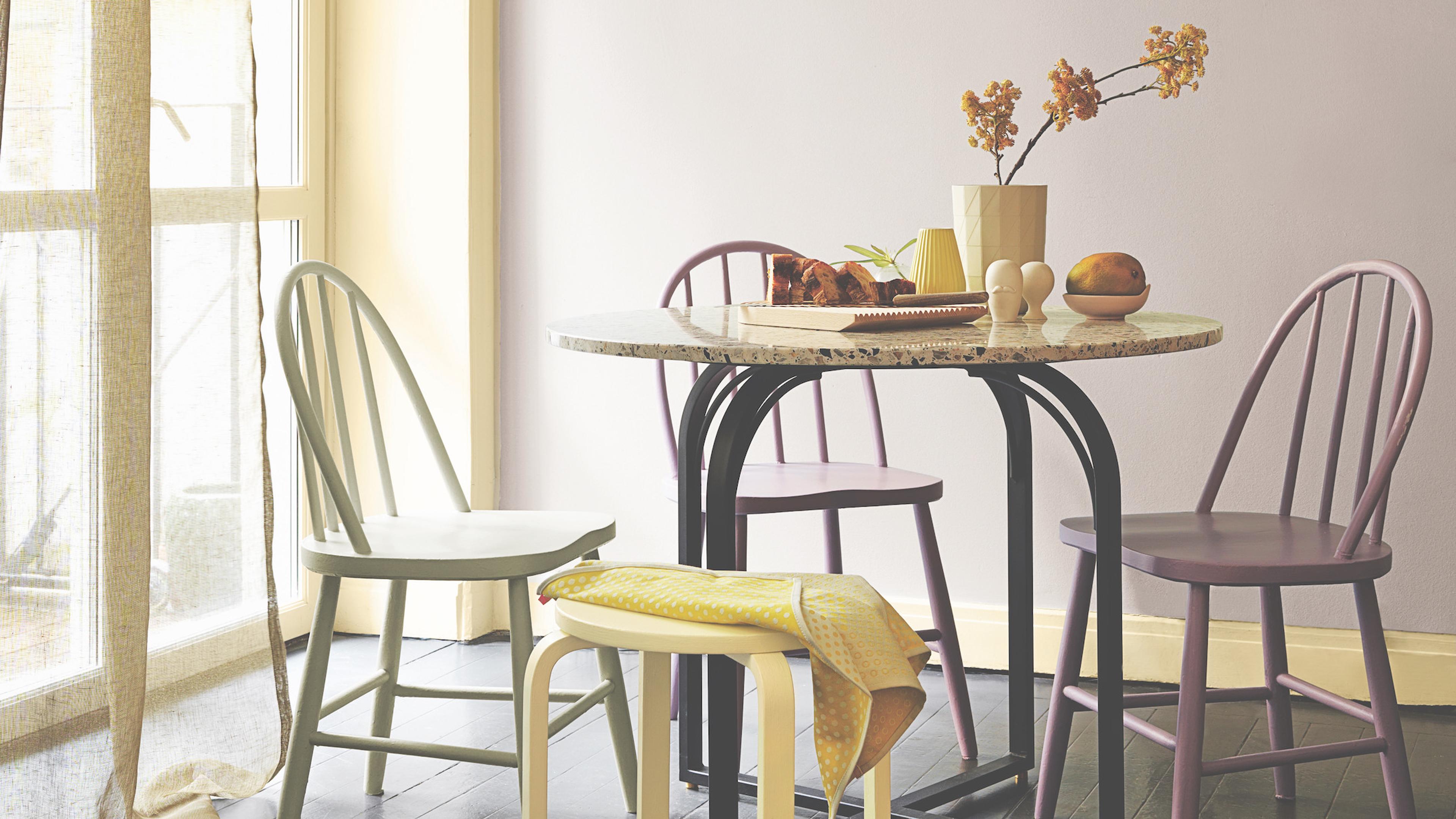 A bright dining room features a round granite table surrounded by colorful chairs and a stool, set with flowers, fruit, and pastries.