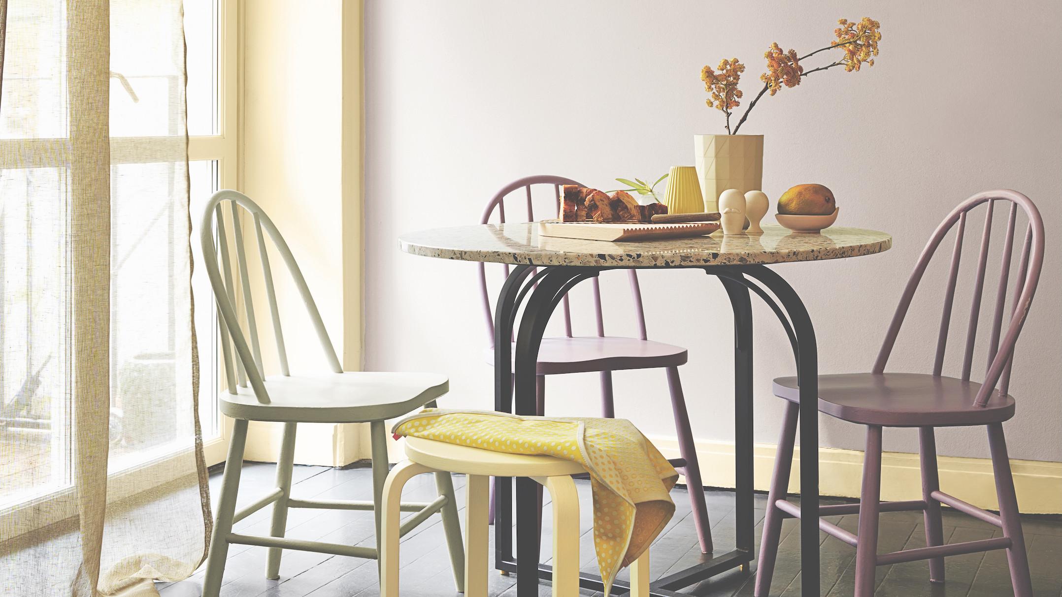 A bright dining room features a round granite table surrounded by colorful chairs and a stool, set with flowers, fruit, and pastries.