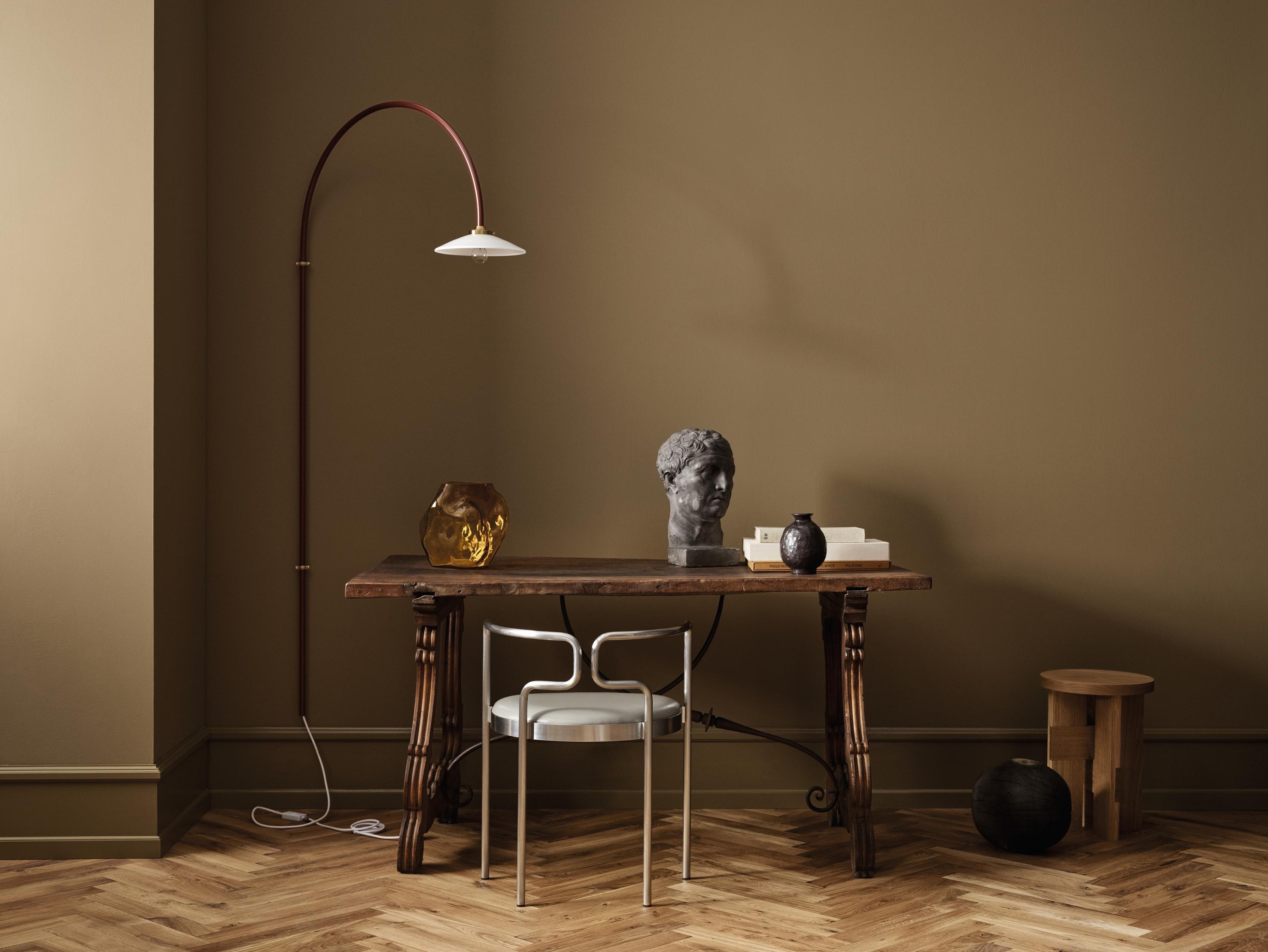 An olive green room with herringbone wood floor, featuring a dark wooden desk, a modern silver chair, an arched floor lamp, a classical bust, and a golden vase.