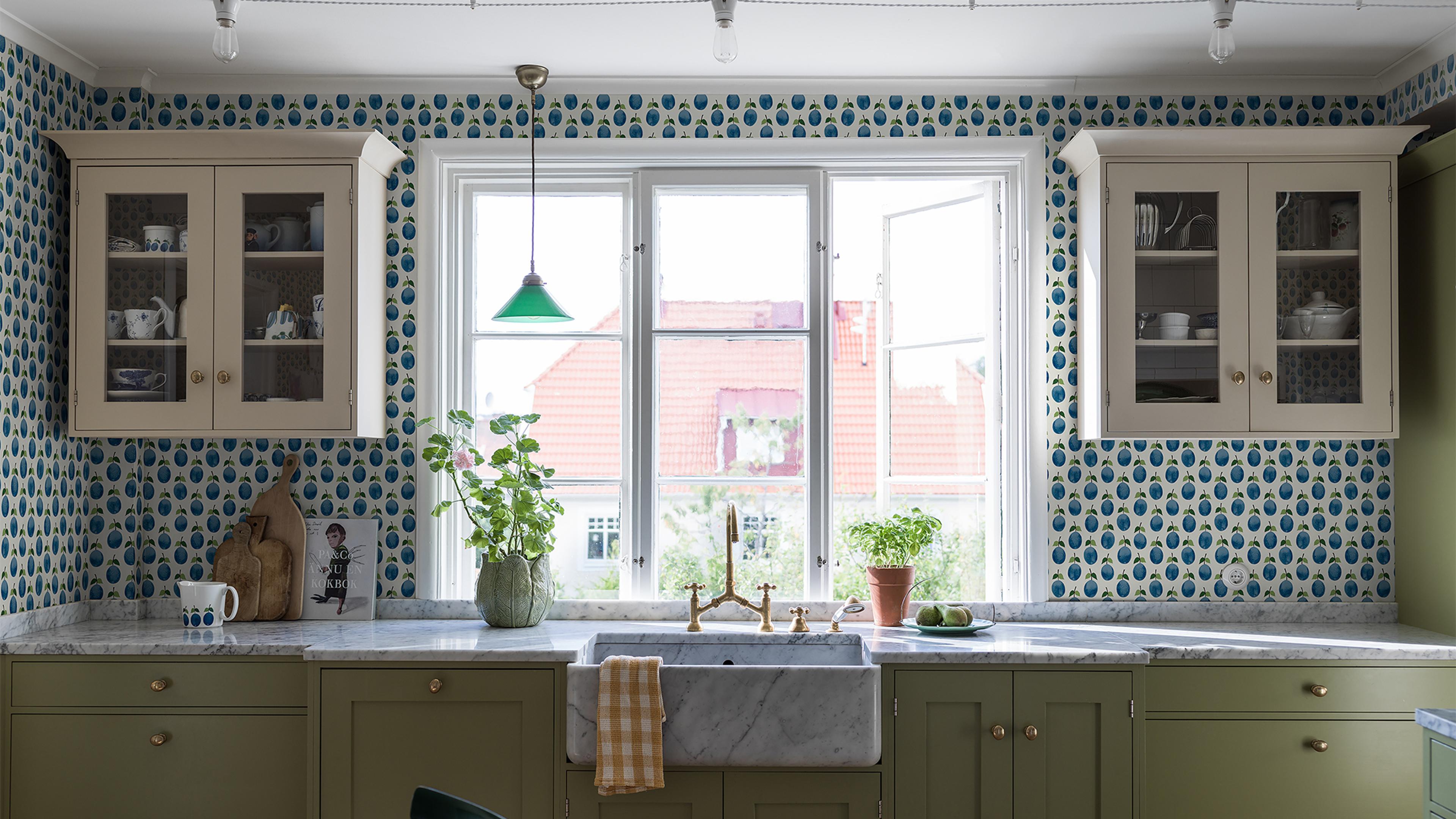 A kitchen with blue and white patterned wallpaper, olive green base cabinets, a white marble counter, and a farmhouse sink under a window with a green pendant light.
