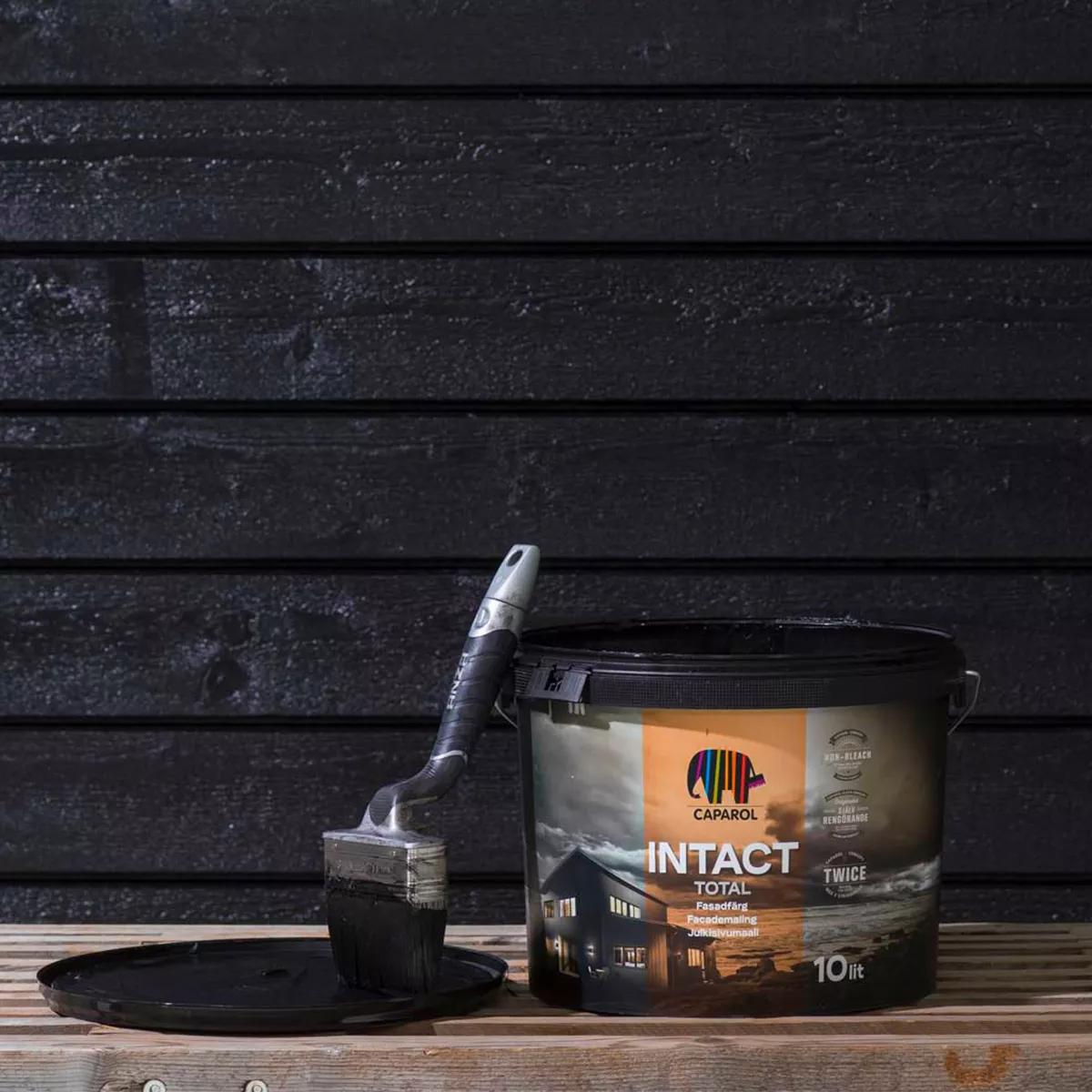 A bucket of black paint and a paint-covered brush sit on a wooden bench against a black wooden wall.