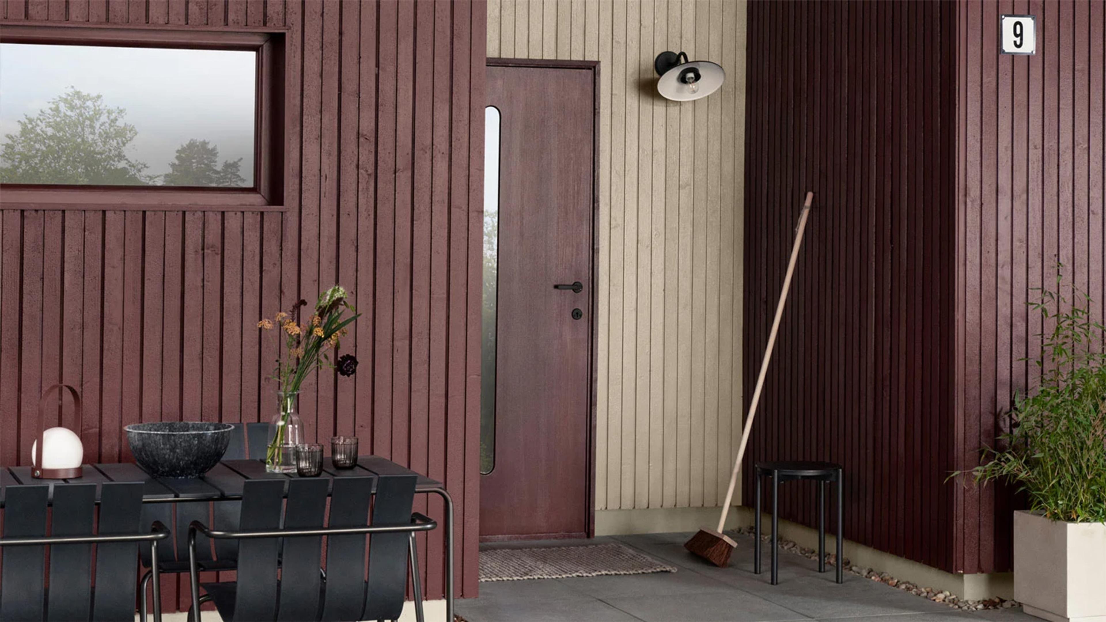 A modern house exterior with vertical reddish-brown and light beige wood siding, an outdoor dining table, a front door, and a broom.