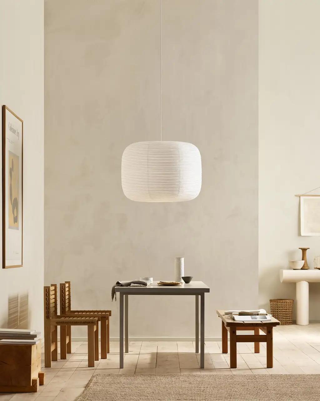 A minimalist room with a large rectangular white paper lantern hanging above a gray table, two wooden chairs, and a wooden bench.