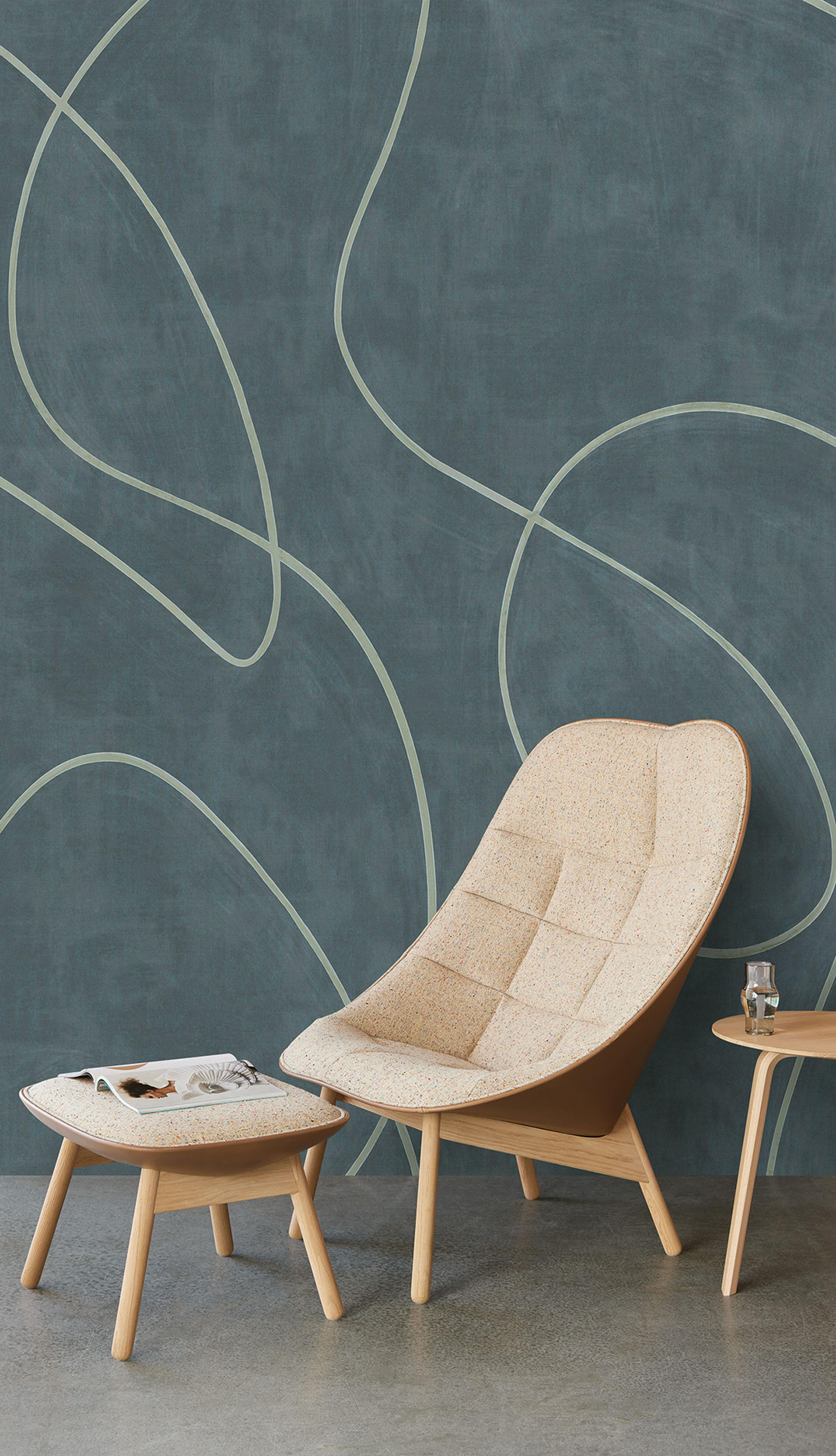 A beige lounge chair and ottoman in a modern room with a teal wall featuring light, wavy abstract lines, and a small wooden side table.