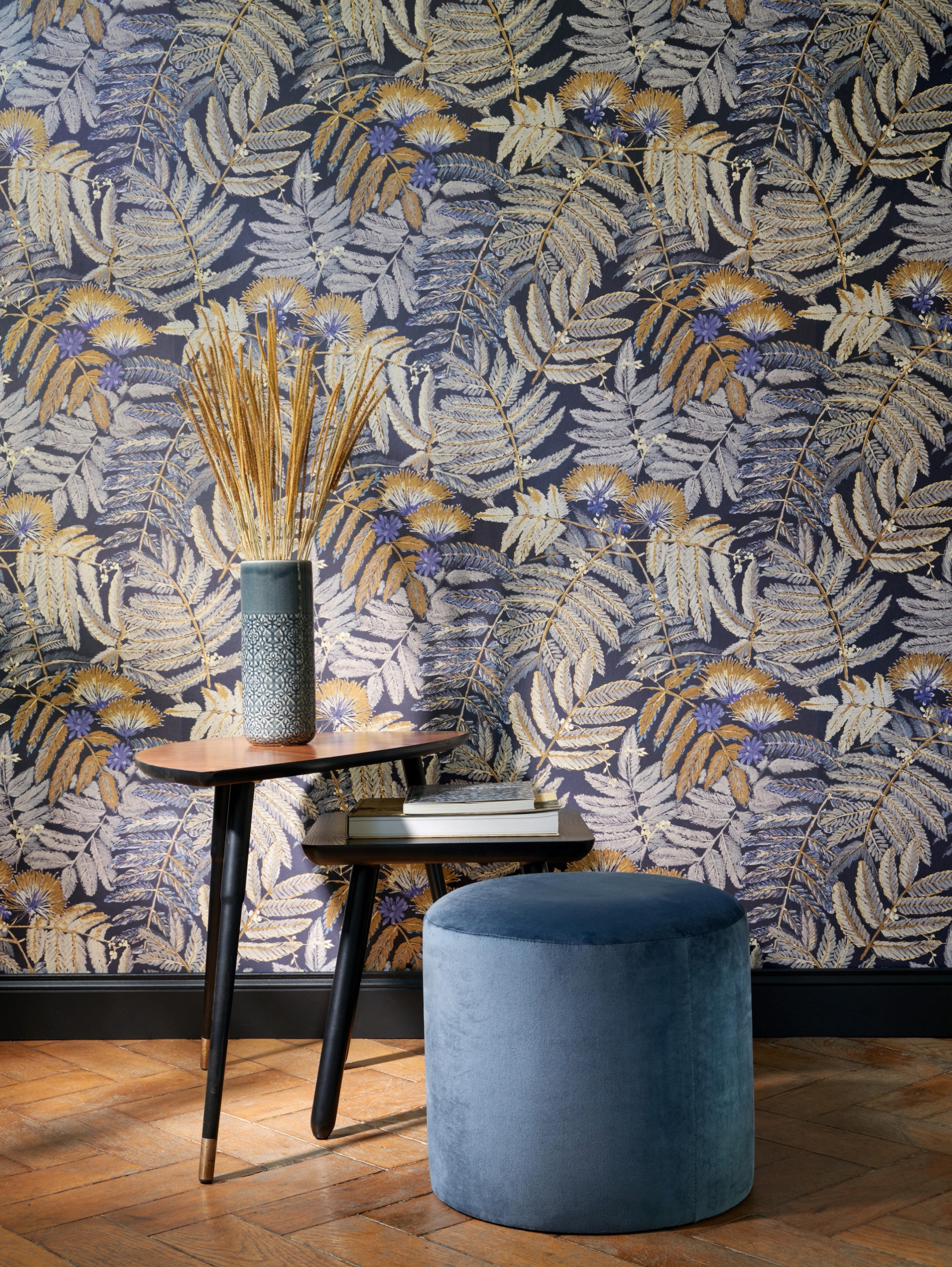 A room corner with dark blue wallpaper featuring gold and purple fern patterns, two wooden side tables, a vase of dried grass, and a round blue velvet ottoman.