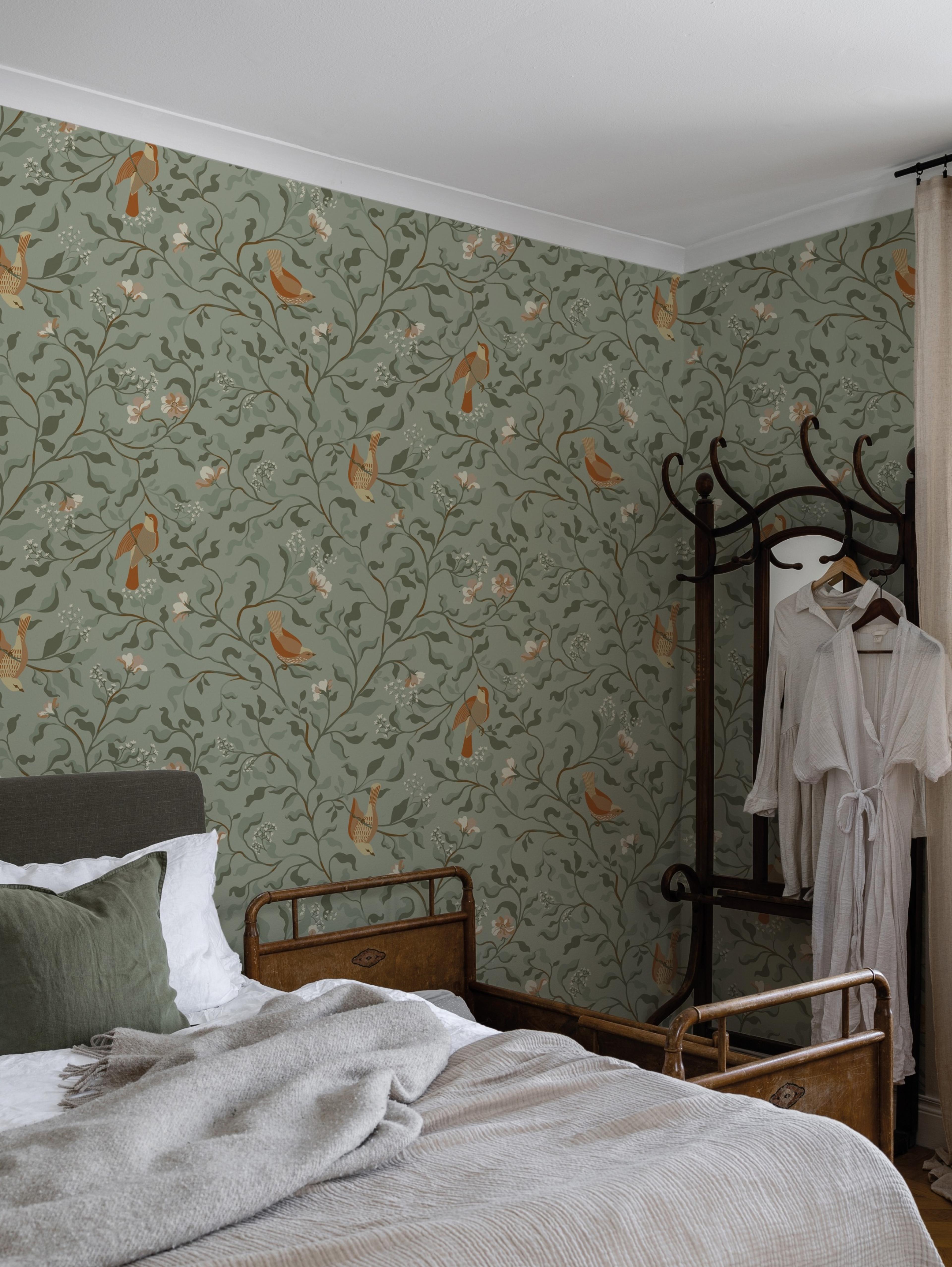 A bedroom with green bird-and-vine patterned wallpaper, a bed with a grey blanket, and a dark wood coat rack.