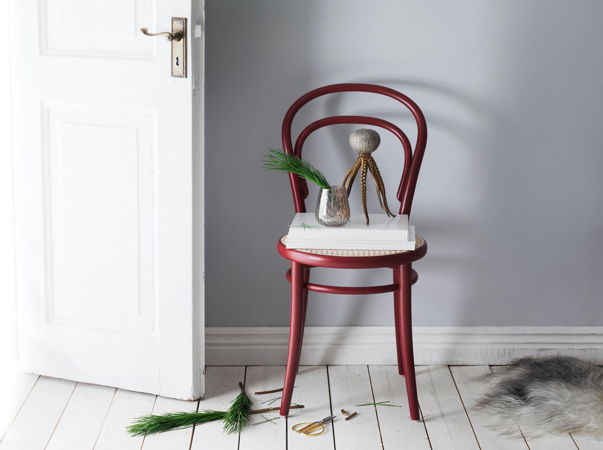 A red wooden chair holds white books, a vase with green branches, and an octopus figurine, placed by an open white door with scattered greenery and scissors on the floor.