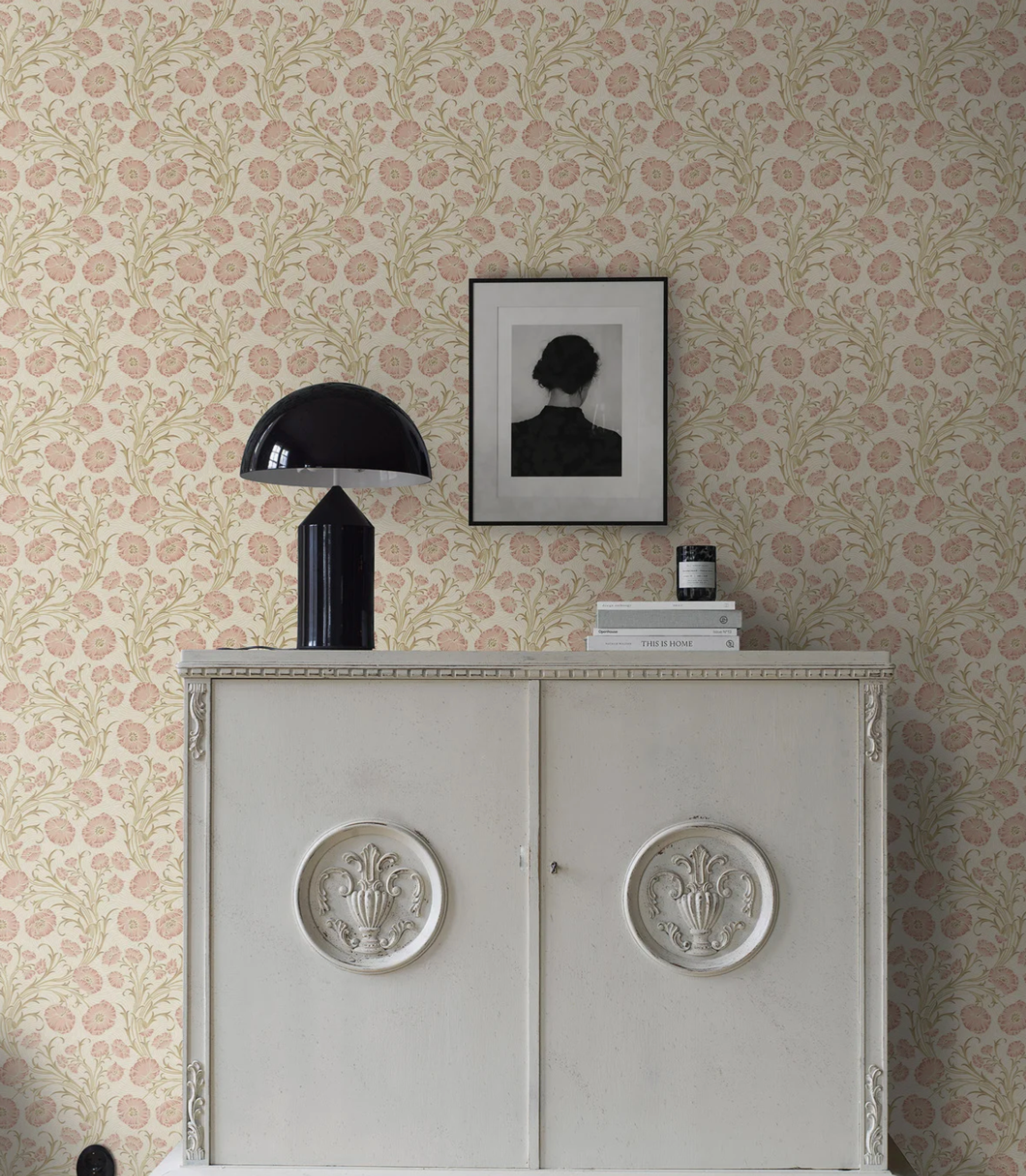 Elegant interior with pink floral wallpaper, a white decorative cabinet, a black table lamp, and framed art.