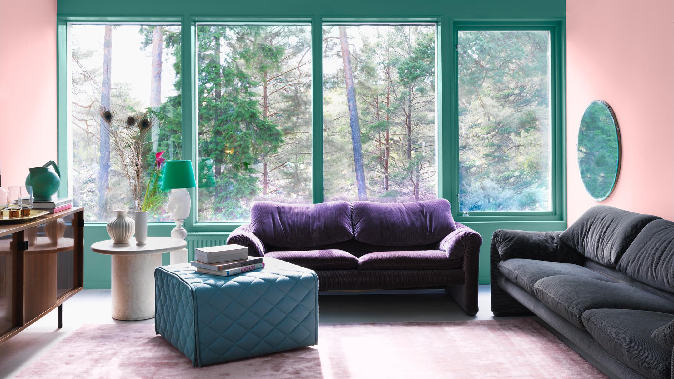 A modern living room with pink walls, teal window frames, a forest view through large windows, purple and dark gray sofas, and a teal ottoman.