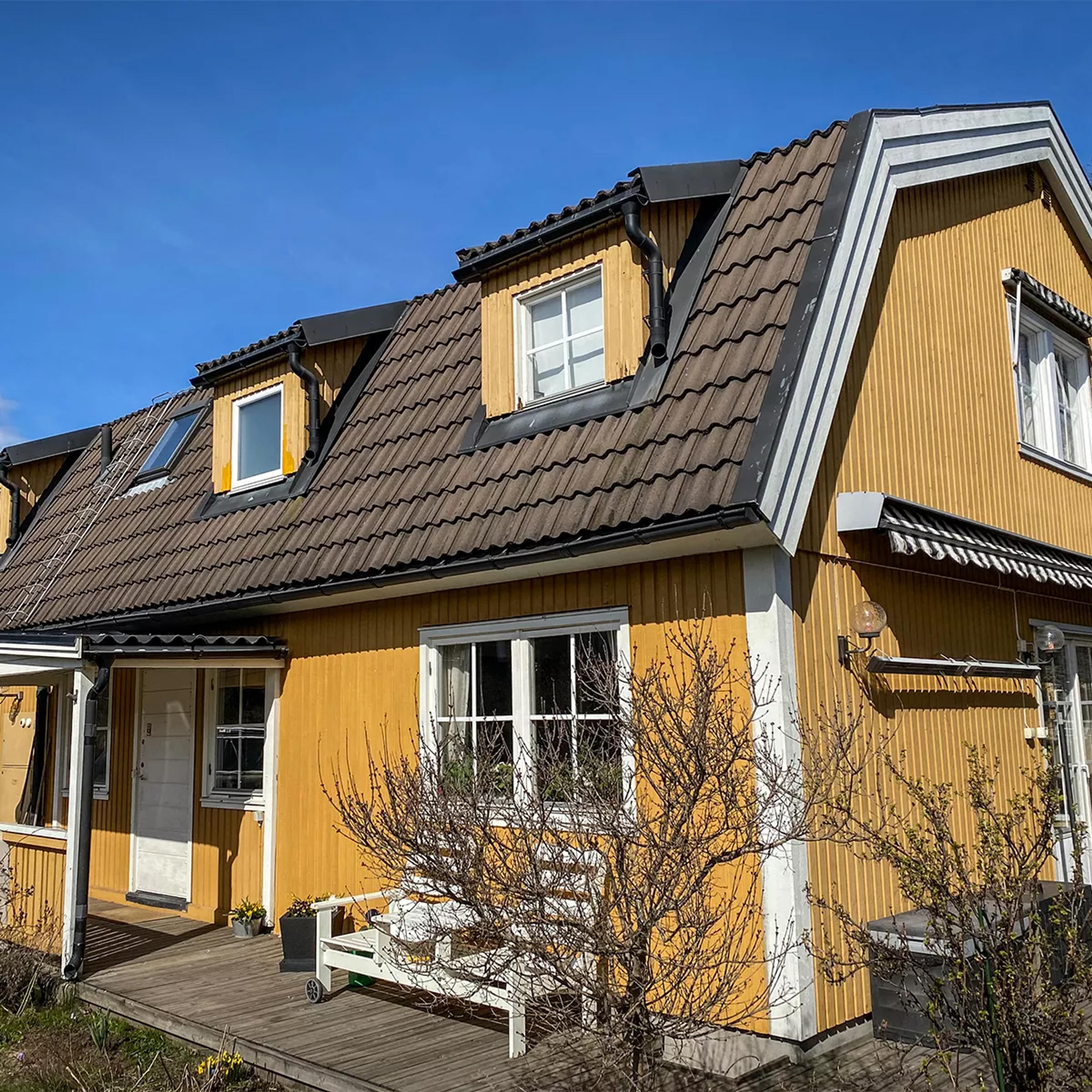 A yellow house with a brown tiled roof, dormer windows, and a wooden deck, under a clear blue sky.