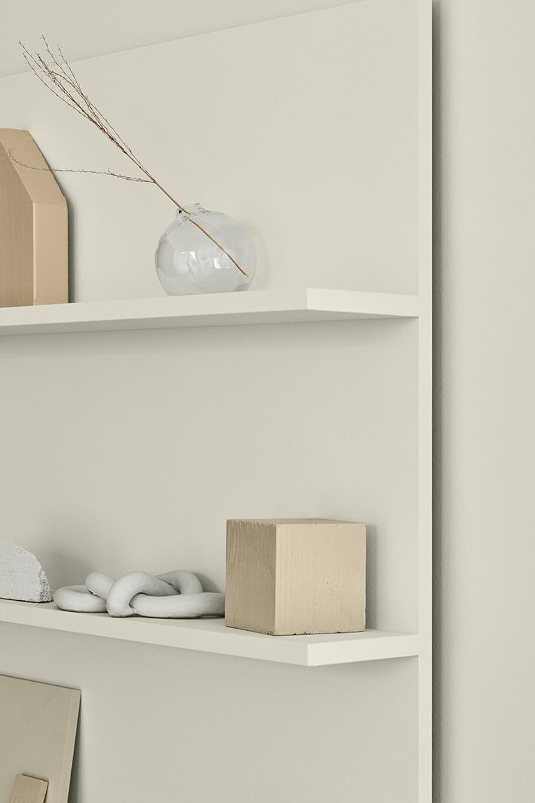 Two light-colored shelves display minimalist decor: a clear vase with a dried branch, a beige house, a beige cube, and a white decorative knot.
