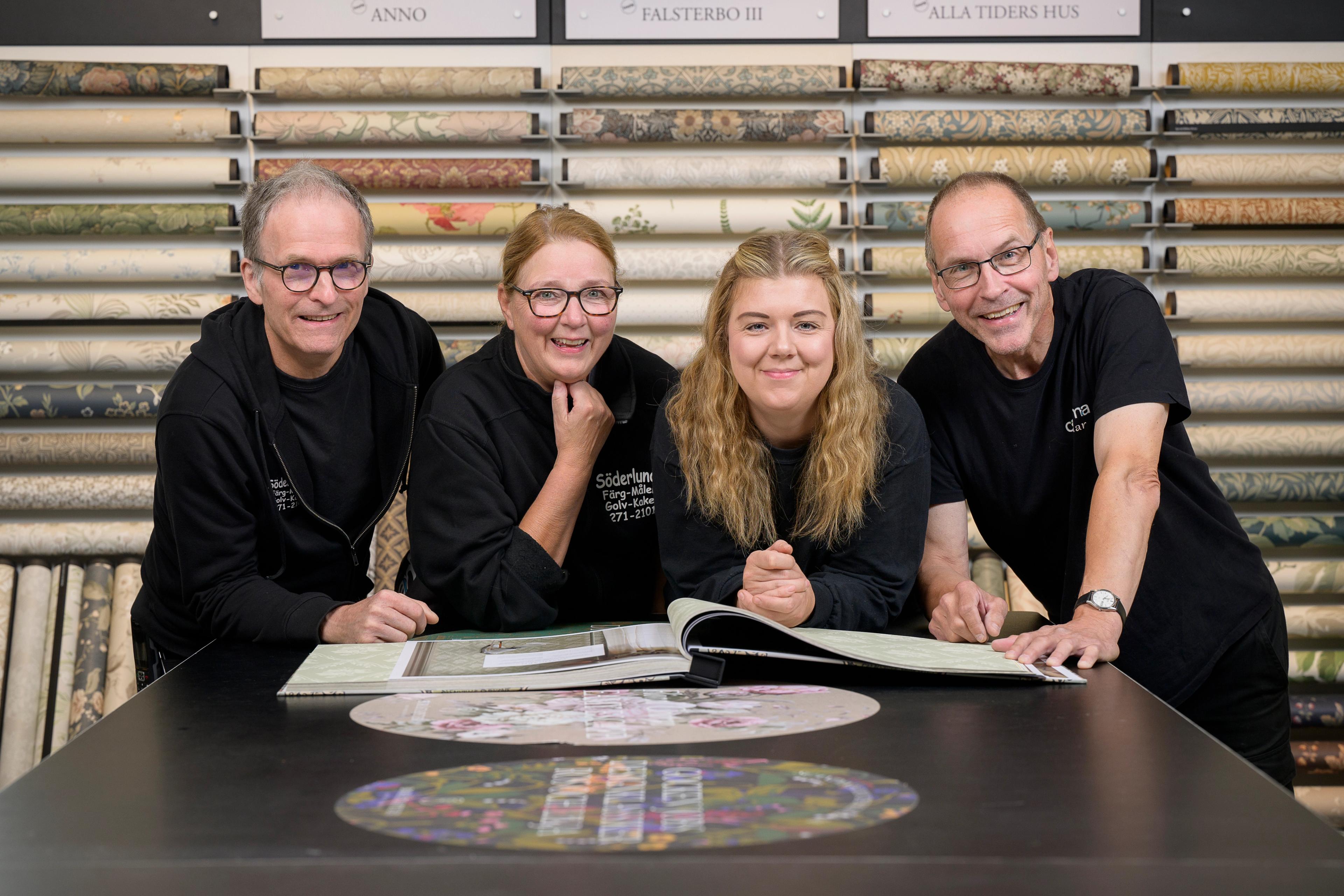 Four smiling people, two men and two women, in a wallpaper store, looking at design books on a table with rolls of wallpaper behind them.