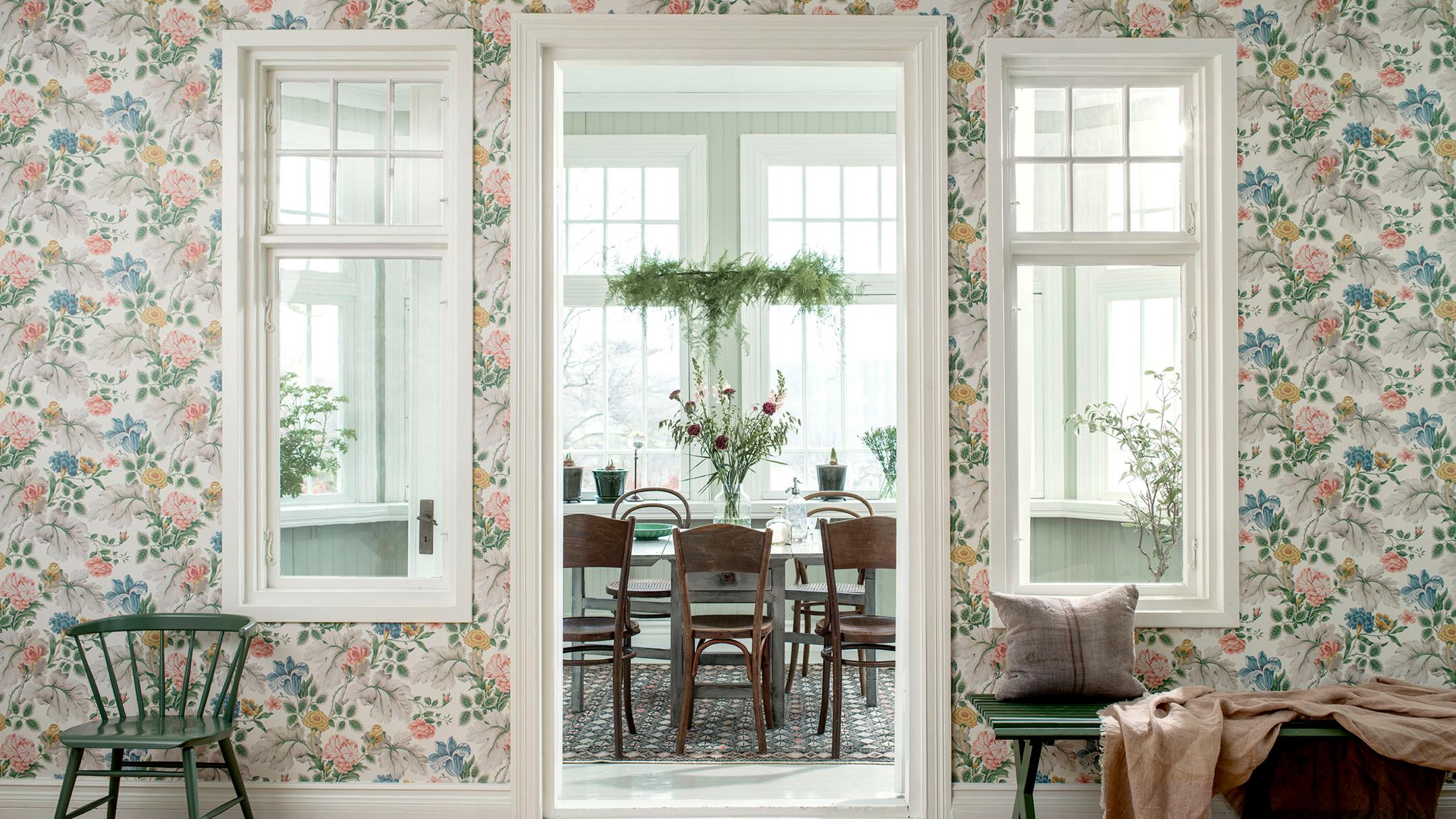 A room with light floral wallpaper, featuring a doorway opening to a dining room, flanked by two windows. A green chair is on the left, and a green bench is on the right.