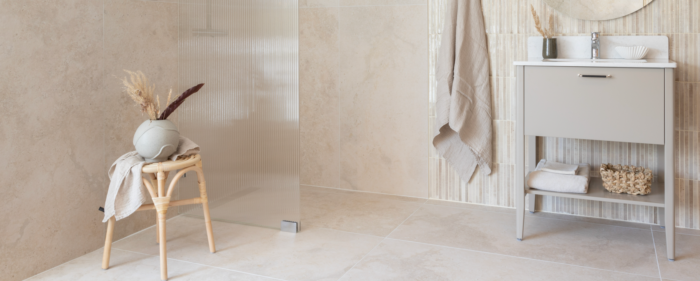 Modern, neutral-toned bathroom featuring a grey vanity with sink, a glass shower partition, and a decorative rattan stool.