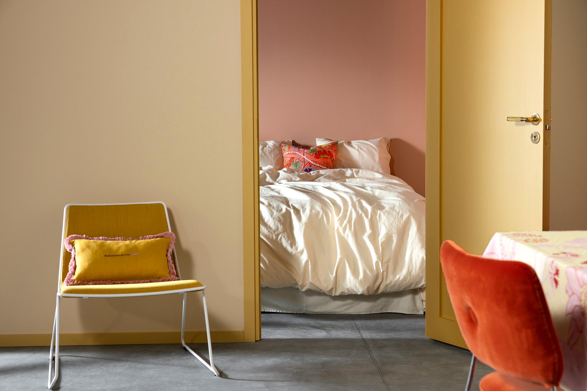 A beige doorway frames a pink-walled bedroom with a bed, while a yellow chair sits in the foreground.