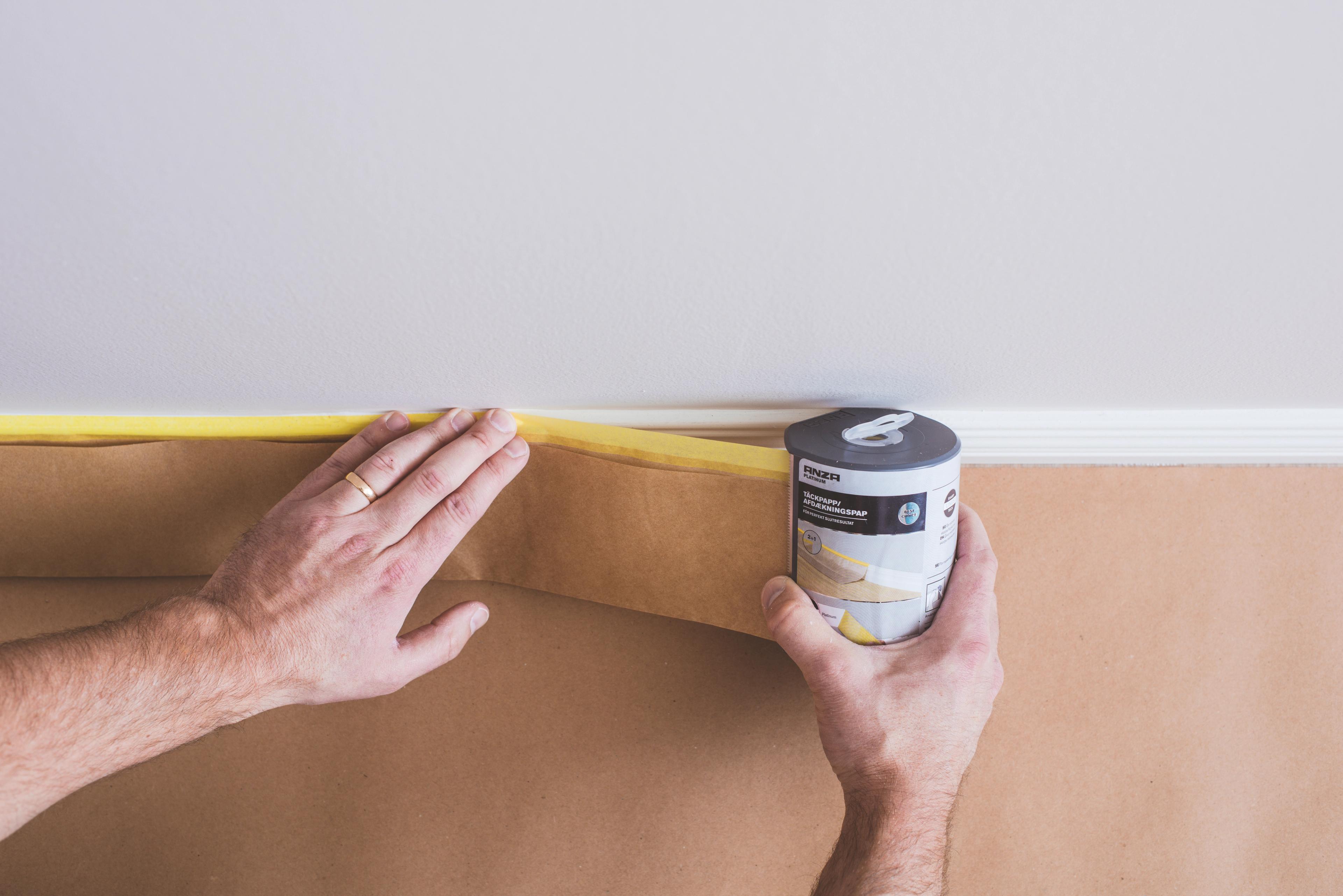 A person applies masking paper with yellow painter's tape along a wall, below a white trim.