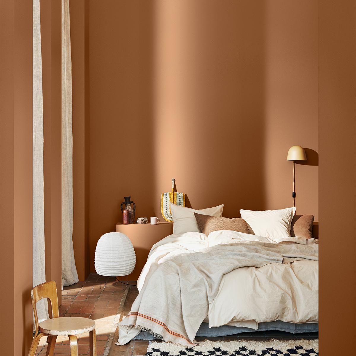 A warm, minimalist bedroom with terracotta walls, a bed dressed in cream linen, and a vintage wooden chair, bathed in sunlight.