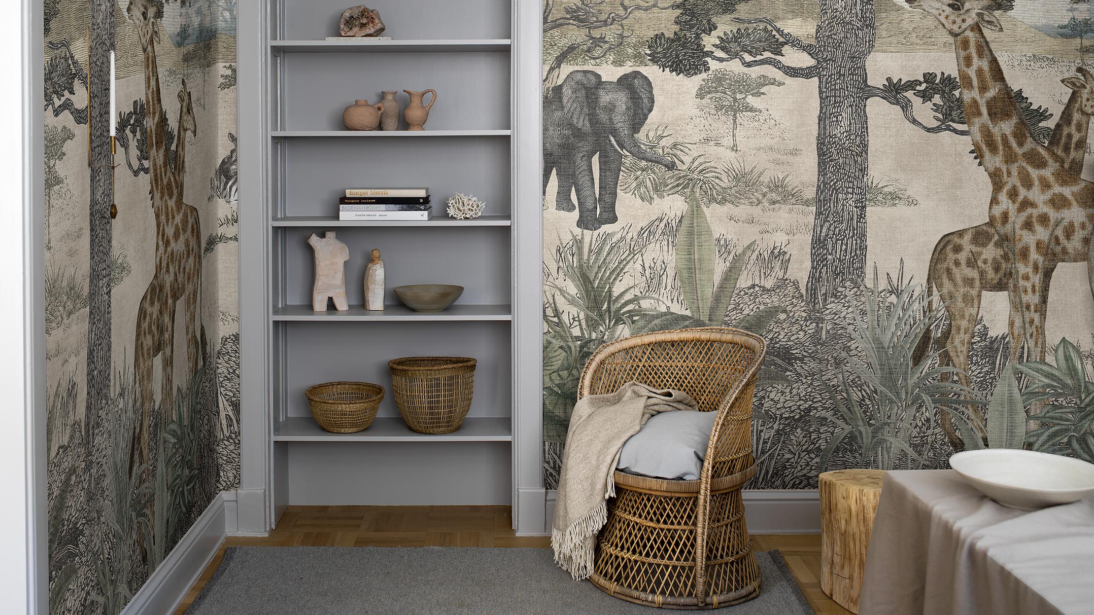 A room with safari-themed wallpaper featuring giraffes and an elephant, a gray built-in bookshelf, and a wicker chair.
