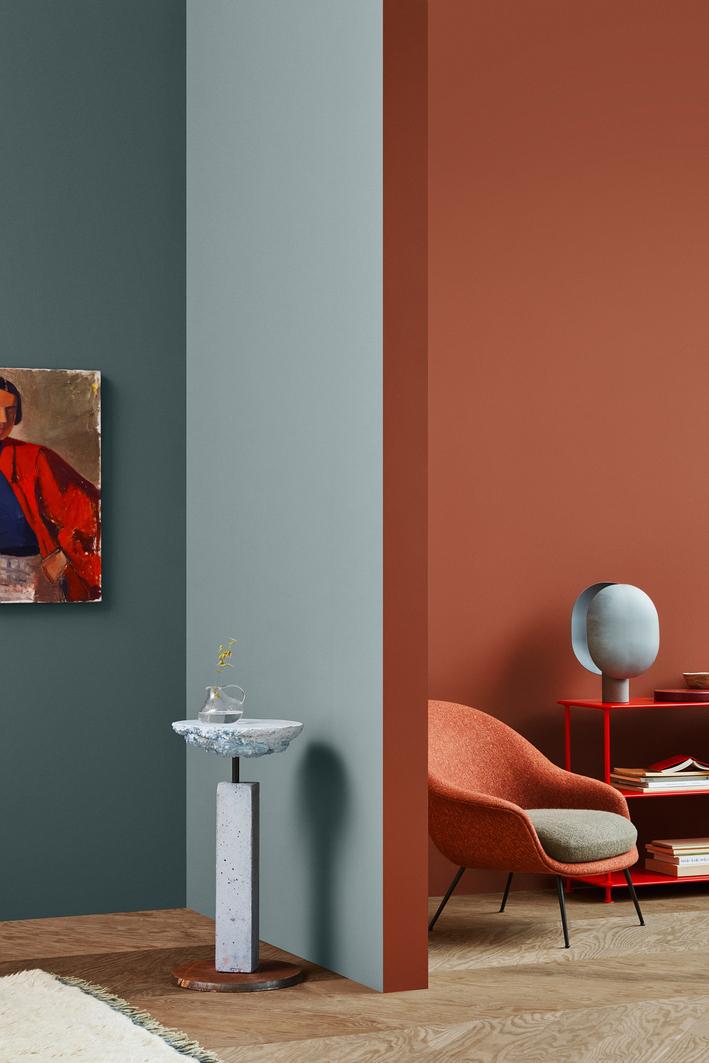 A modern interior with color-blocked walls in teal, light blue, and terracotta, featuring an orange armchair, a concrete pedestal table, and a red shelving unit.