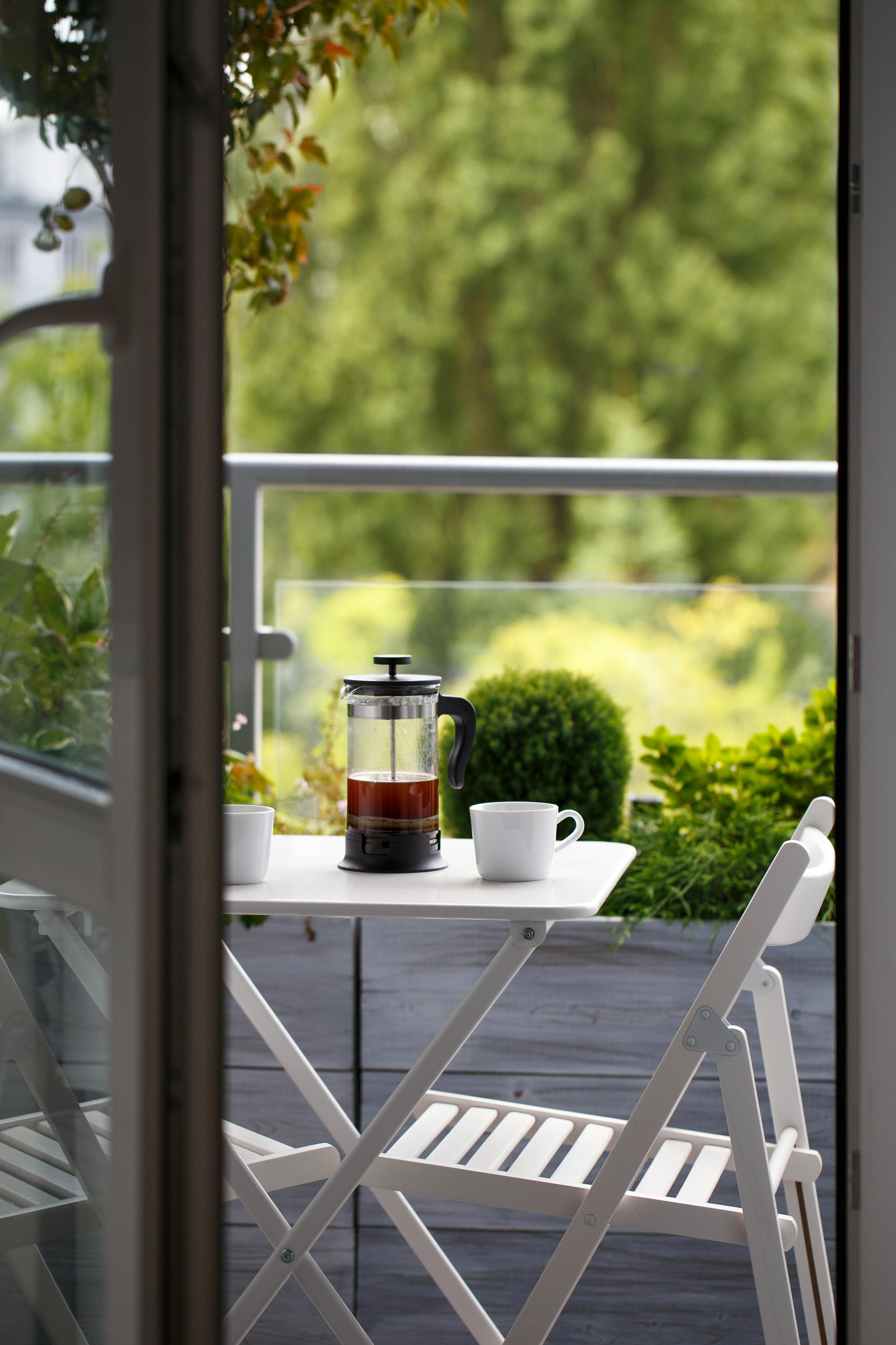 A french press with coffee and two white mugs on a small white balcony table overlooking green foliage.