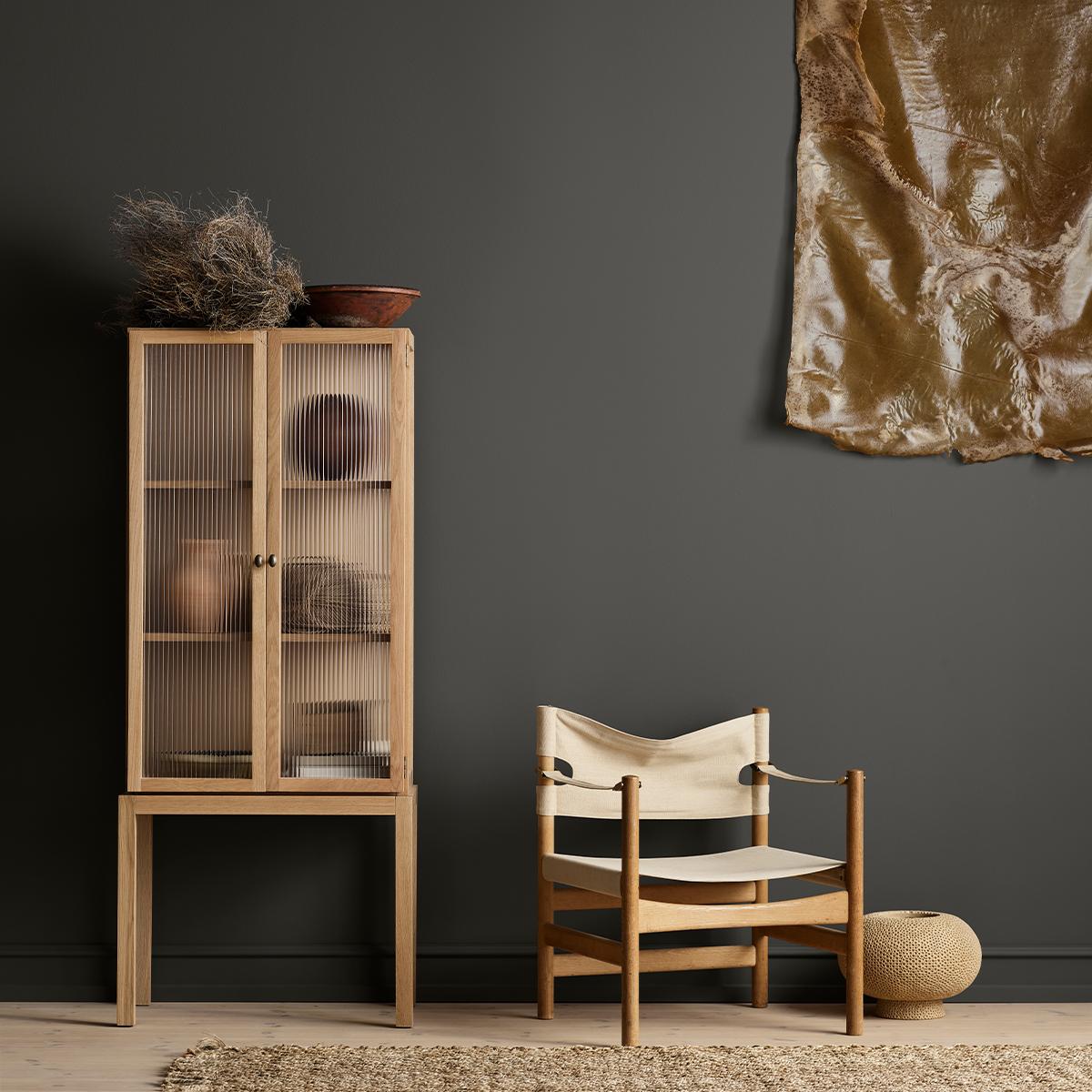 A dark grey room features a light wood cabinet with ribbed glass doors, a canvas and wood chair, and a large golden abstract artwork.