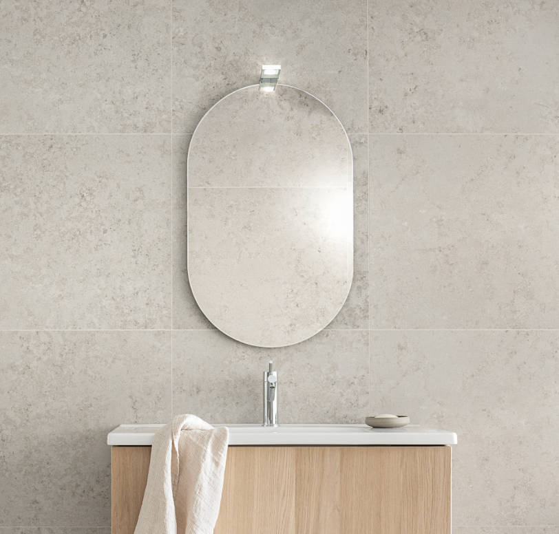 Modern bathroom with an oval mirror, light wood vanity, white sink, and light gray tiled wall.