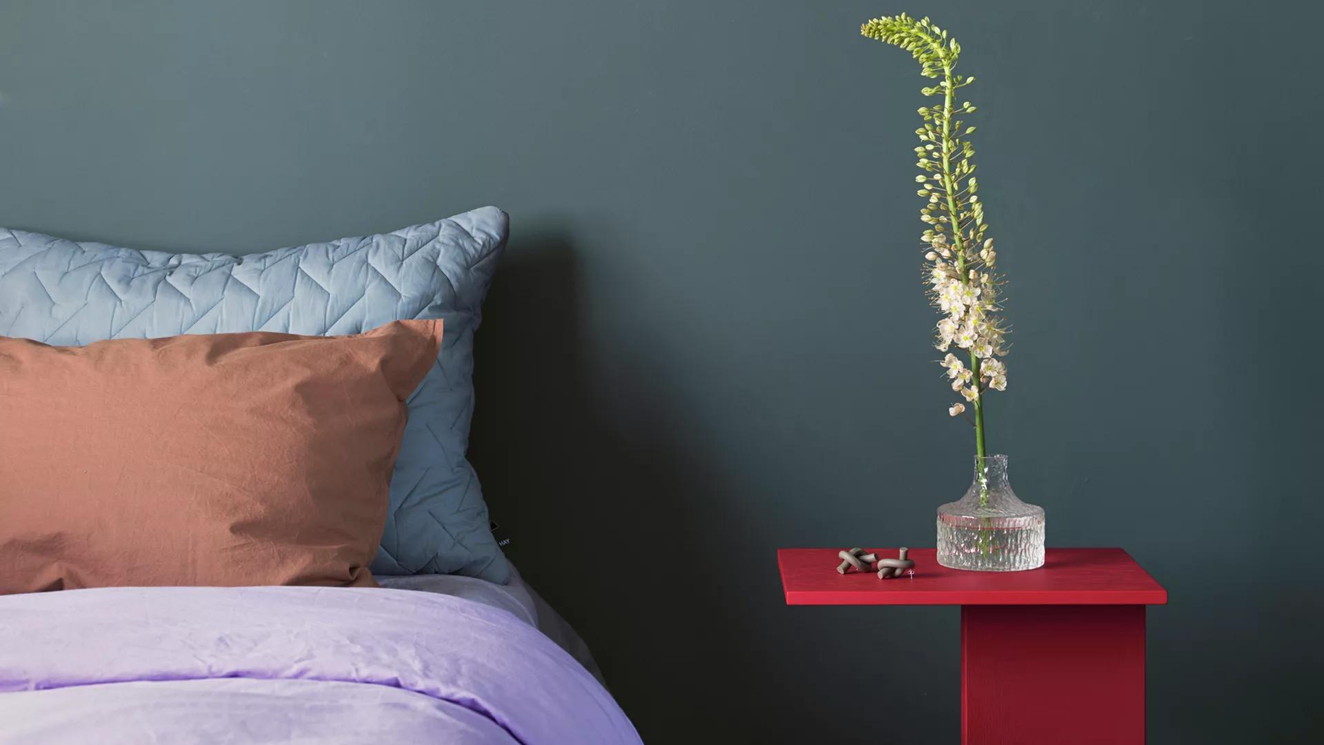 A bed with lavender duvet, blue and peach pillows next to a red side table with a tall white flower in a glass vase against a dark teal wall.
