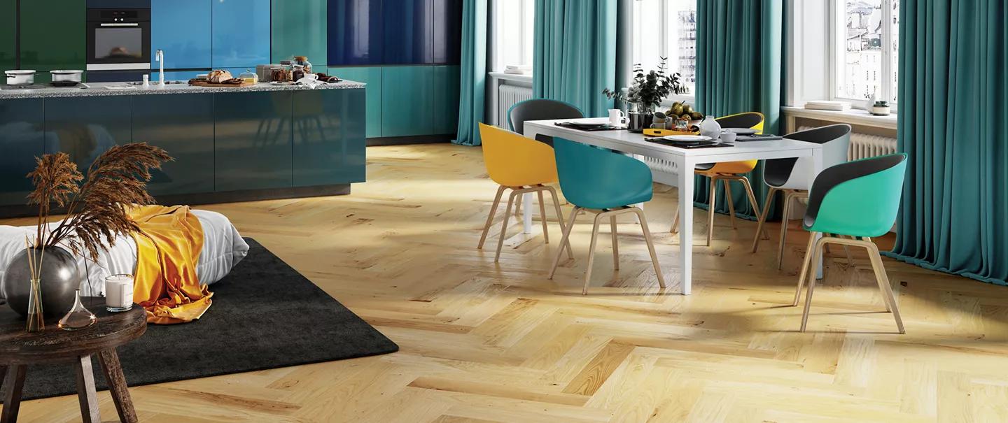 Modern open-plan kitchen and dining area with herringbone wood floor, blue and teal cabinets, and a white dining table with colorful chairs.