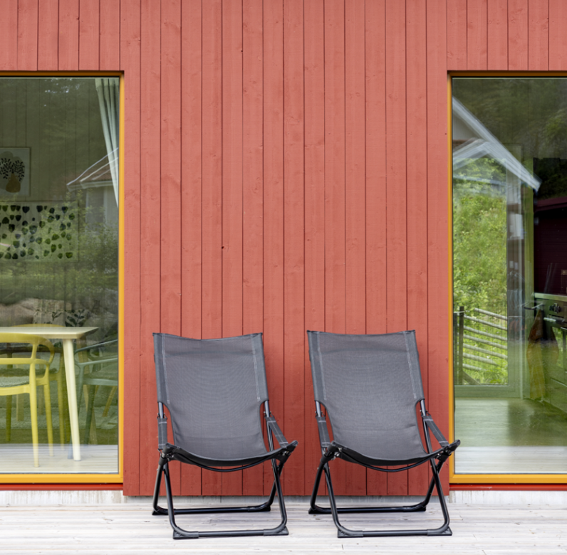 Two dark folding chairs on a wooden deck against a red wooden building with reflective windows.