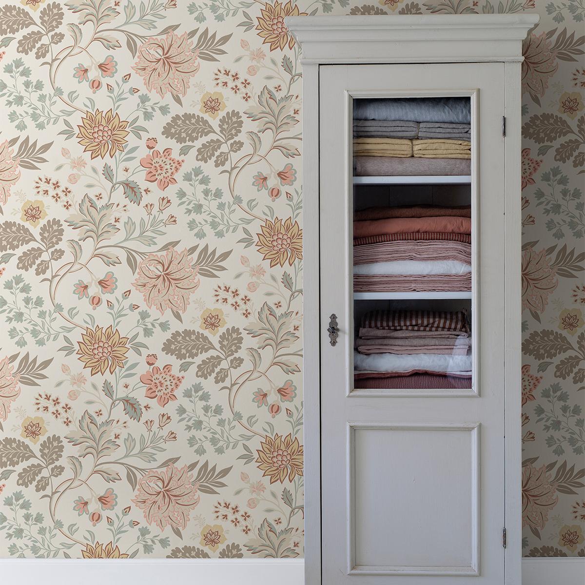 White glass-front cabinet with folded textiles against ornate floral wallpaper.