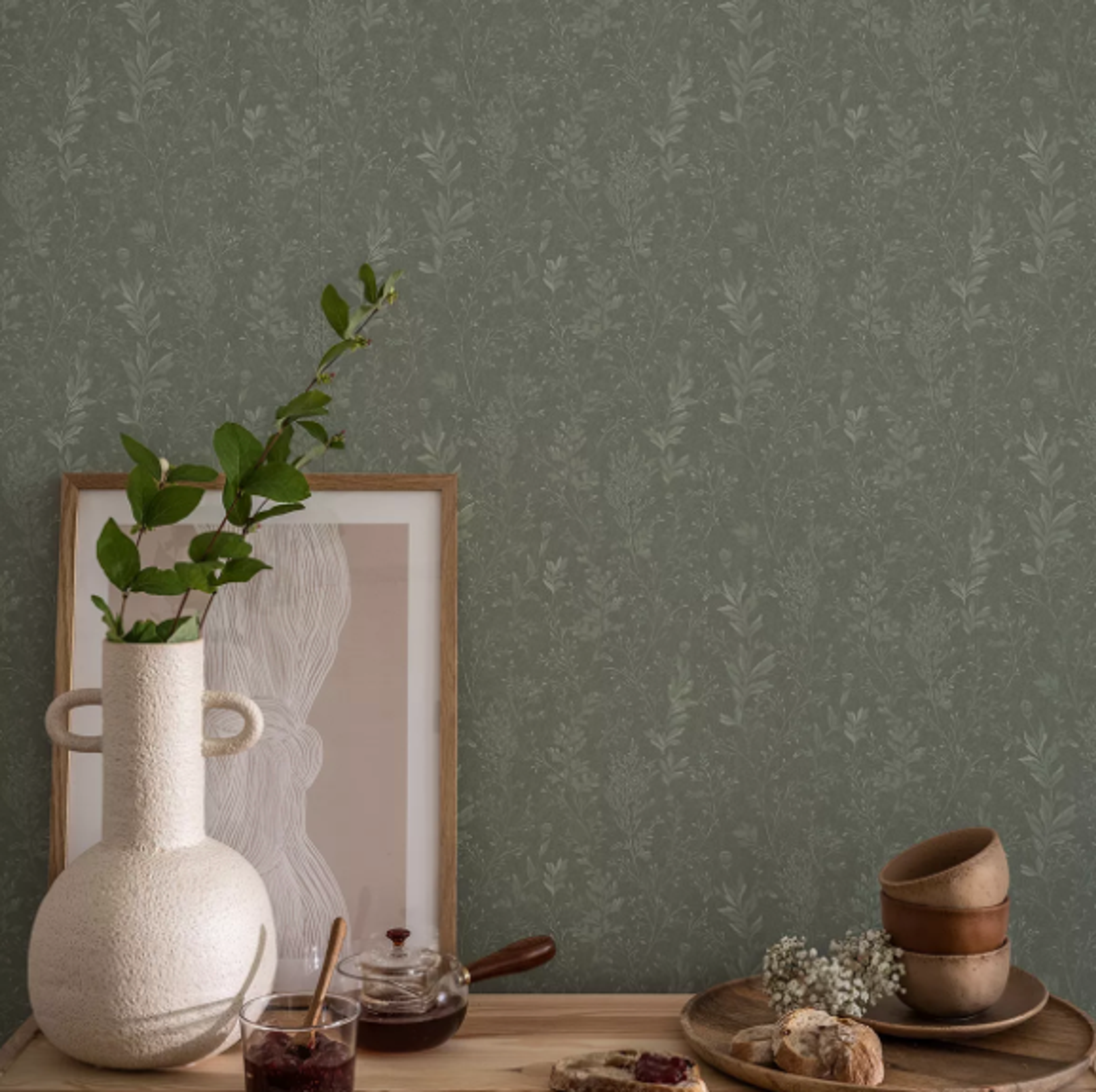 Green wallpaper with a subtle botanical pattern, behind a wooden table with a vase of branches, framed art, jam, a teapot, and a tray with bowls and bread.