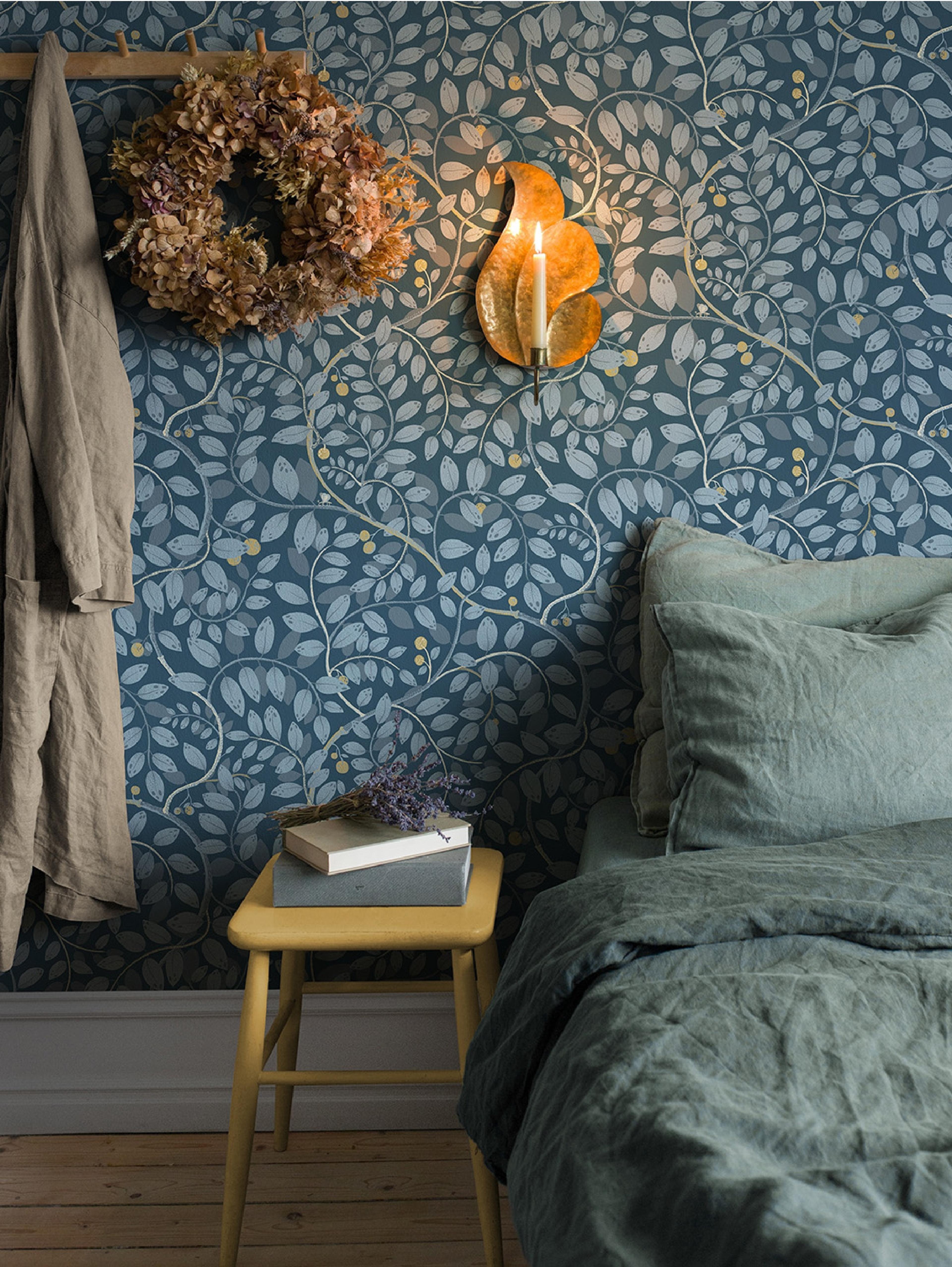 A cozy bedroom features blue wallpaper with a light leaf pattern, a dried wreath, a lit candle sconce, and a bed with sage linen next to a yellow stool holding books and lavender.