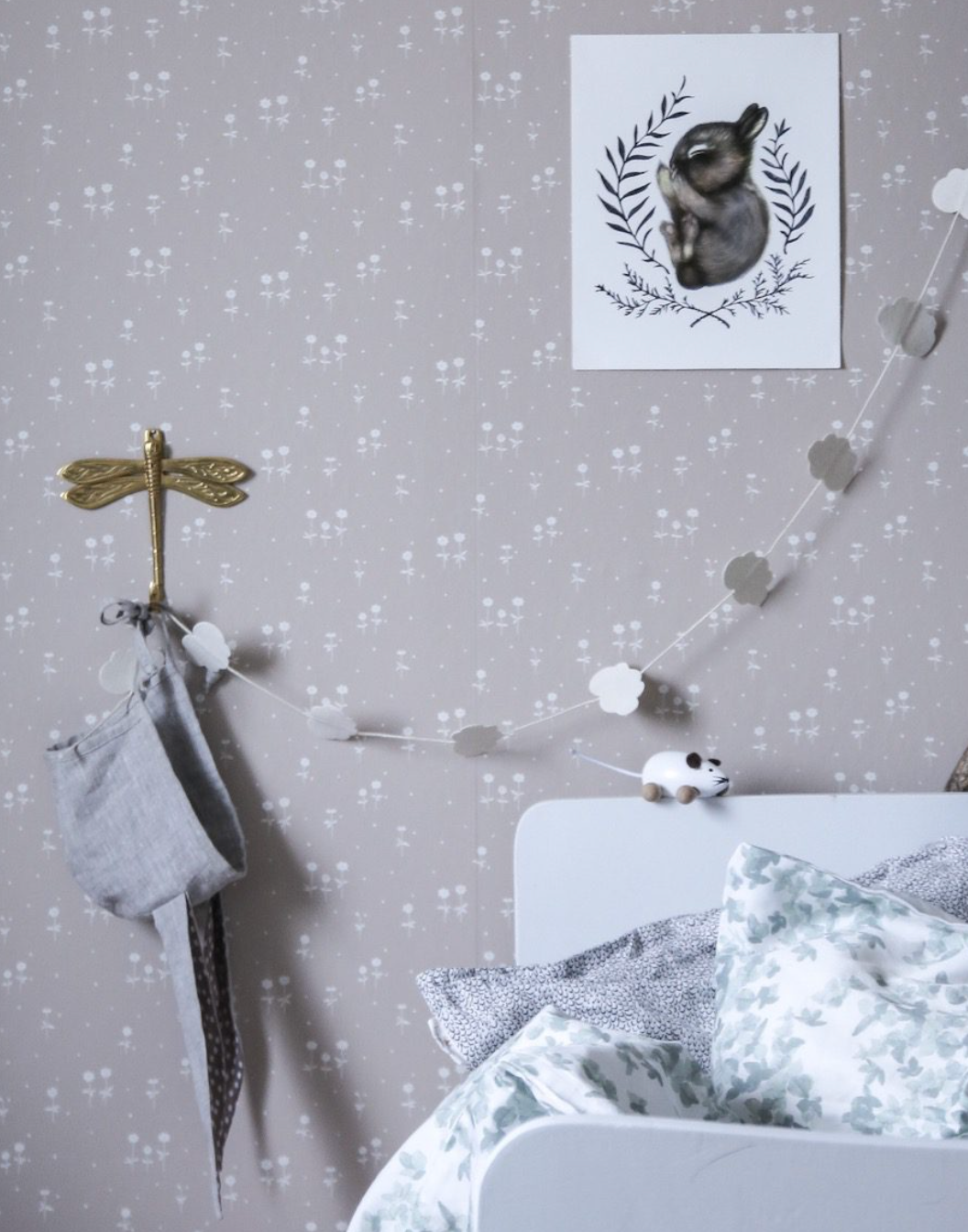 A beige wall with white floral wallpaper, a rabbit art print, a golden dragonfly hook, a white cloud garland, and a toy mouse on a bed.