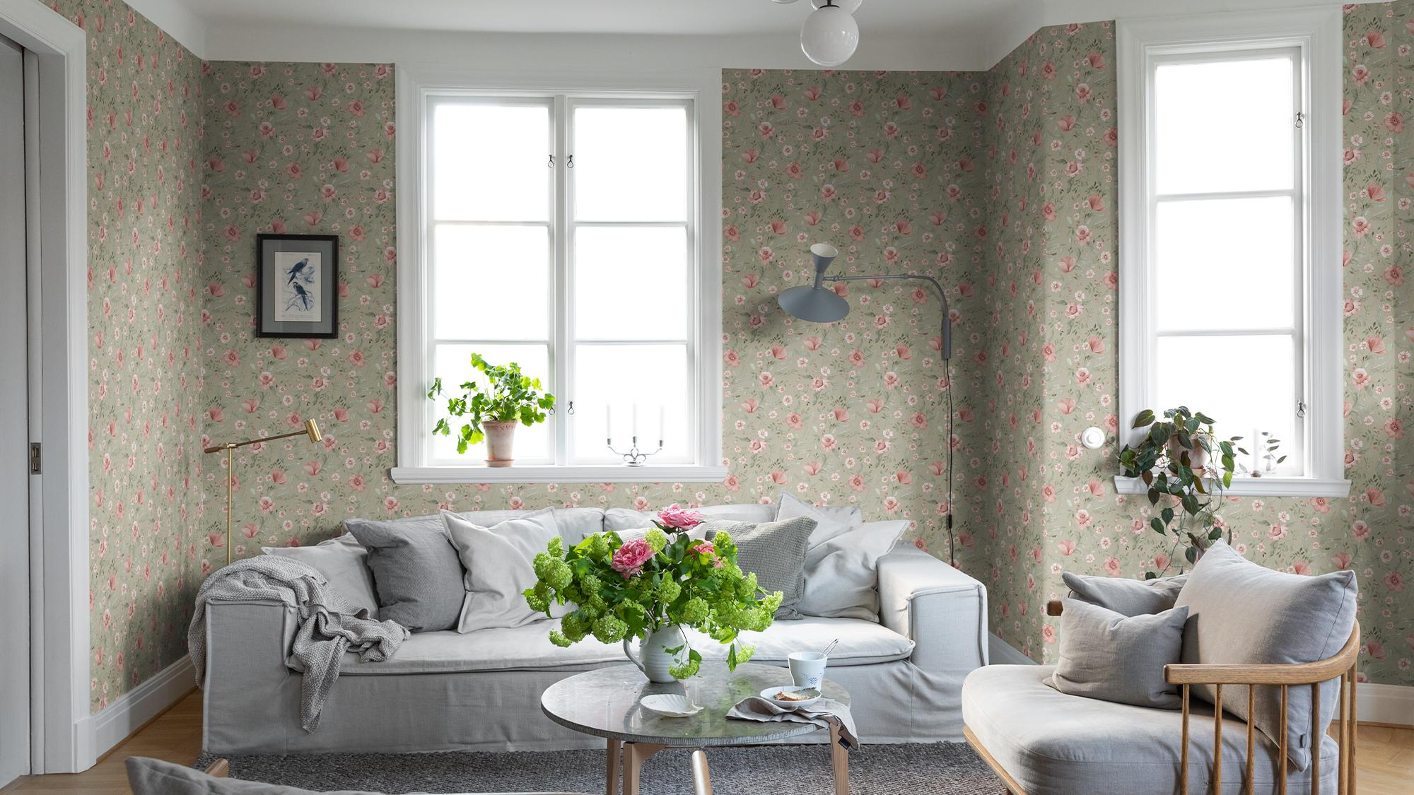 A bright living room with light green floral wallpaper, a grey slipcovered sofa, and a glass coffee table topped with pink flowers.