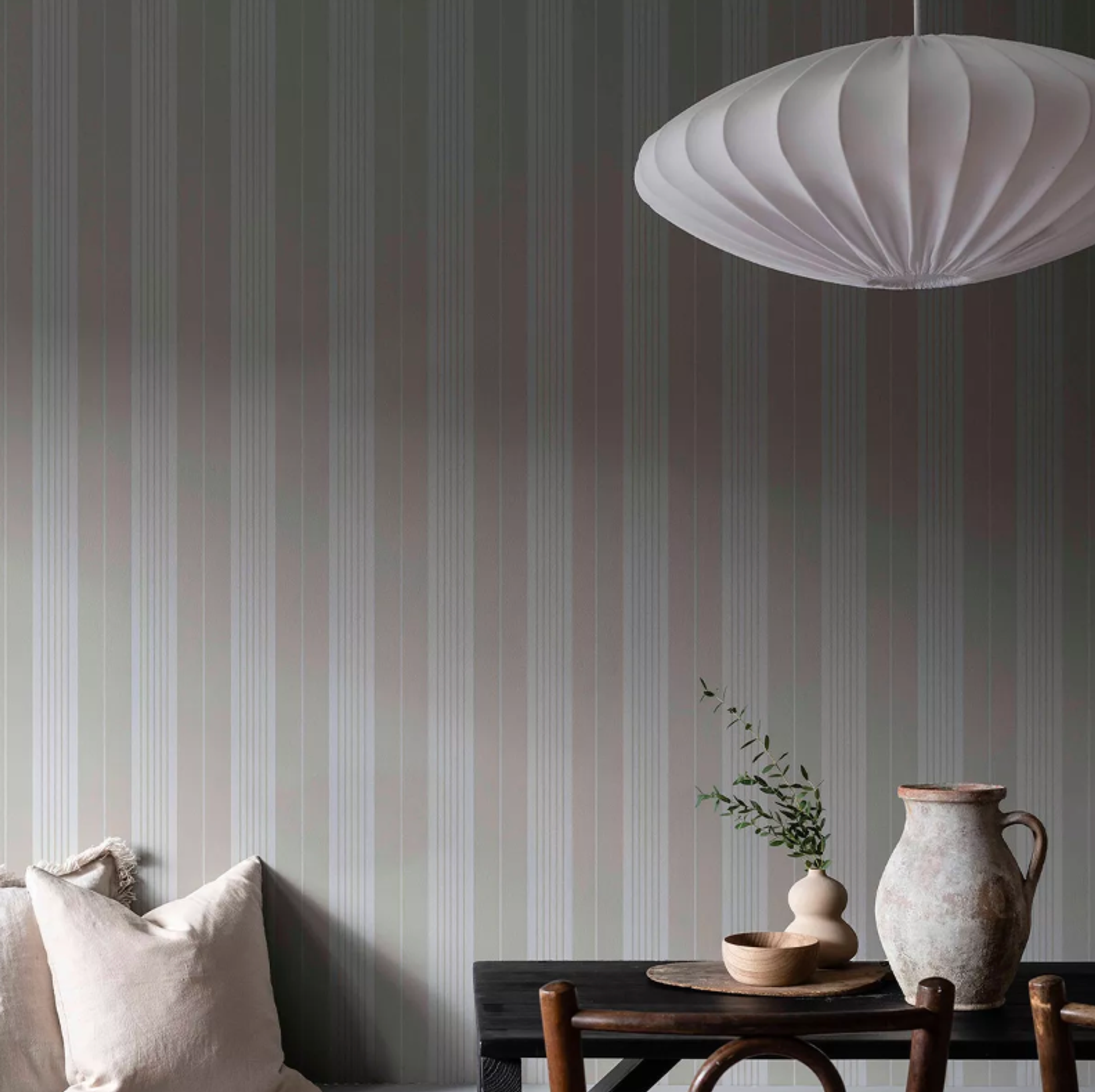 A room with pale green and pink striped wallpaper, a modern white pendant lamp, and a dark wooden table with rustic ceramics and greenery.