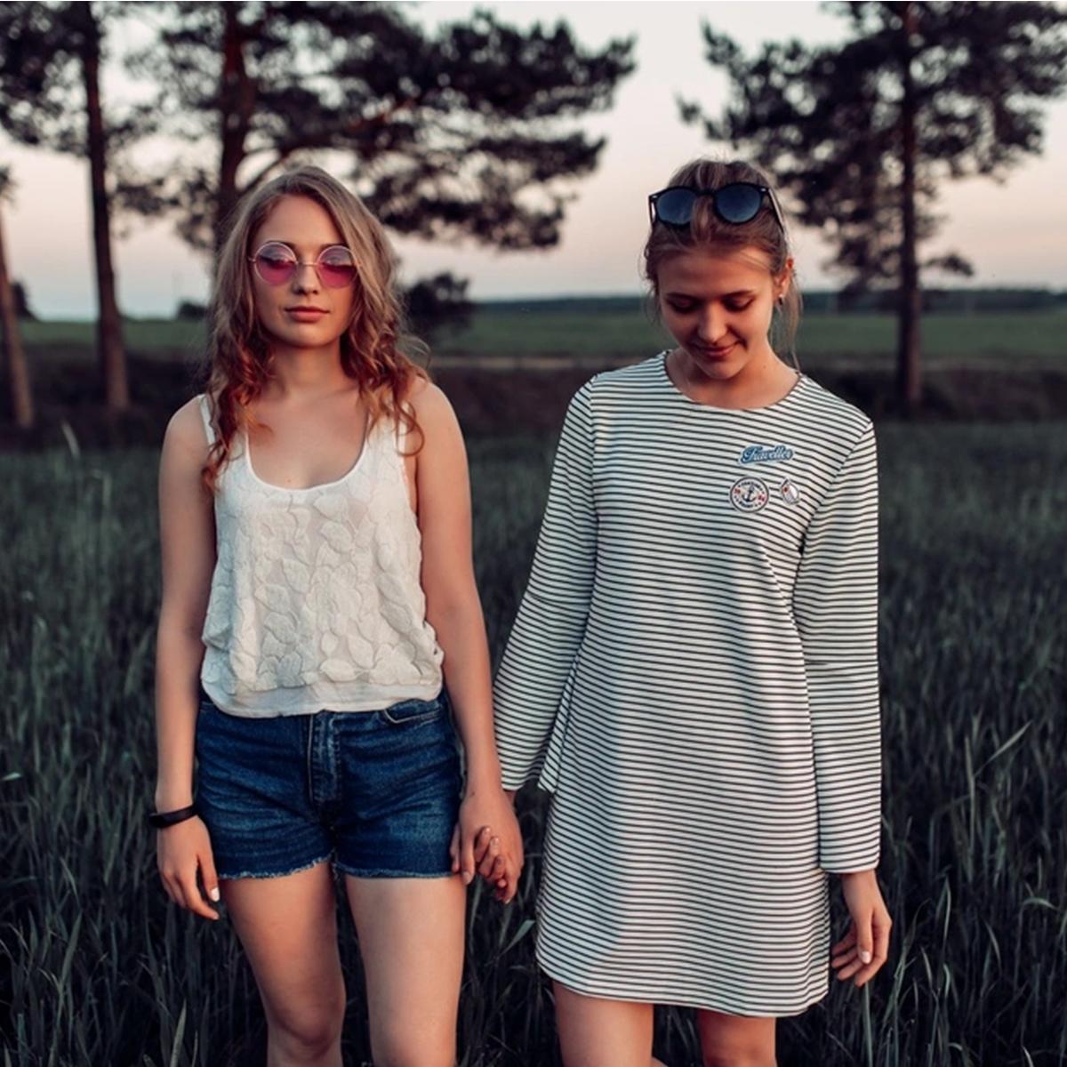 Two young women holding hands in a grassy field with trees in the background.