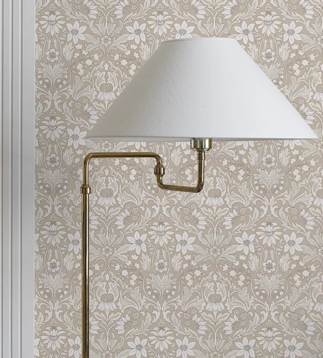Brass lamp with a white shade in front of beige wallpaper featuring a white floral pattern.