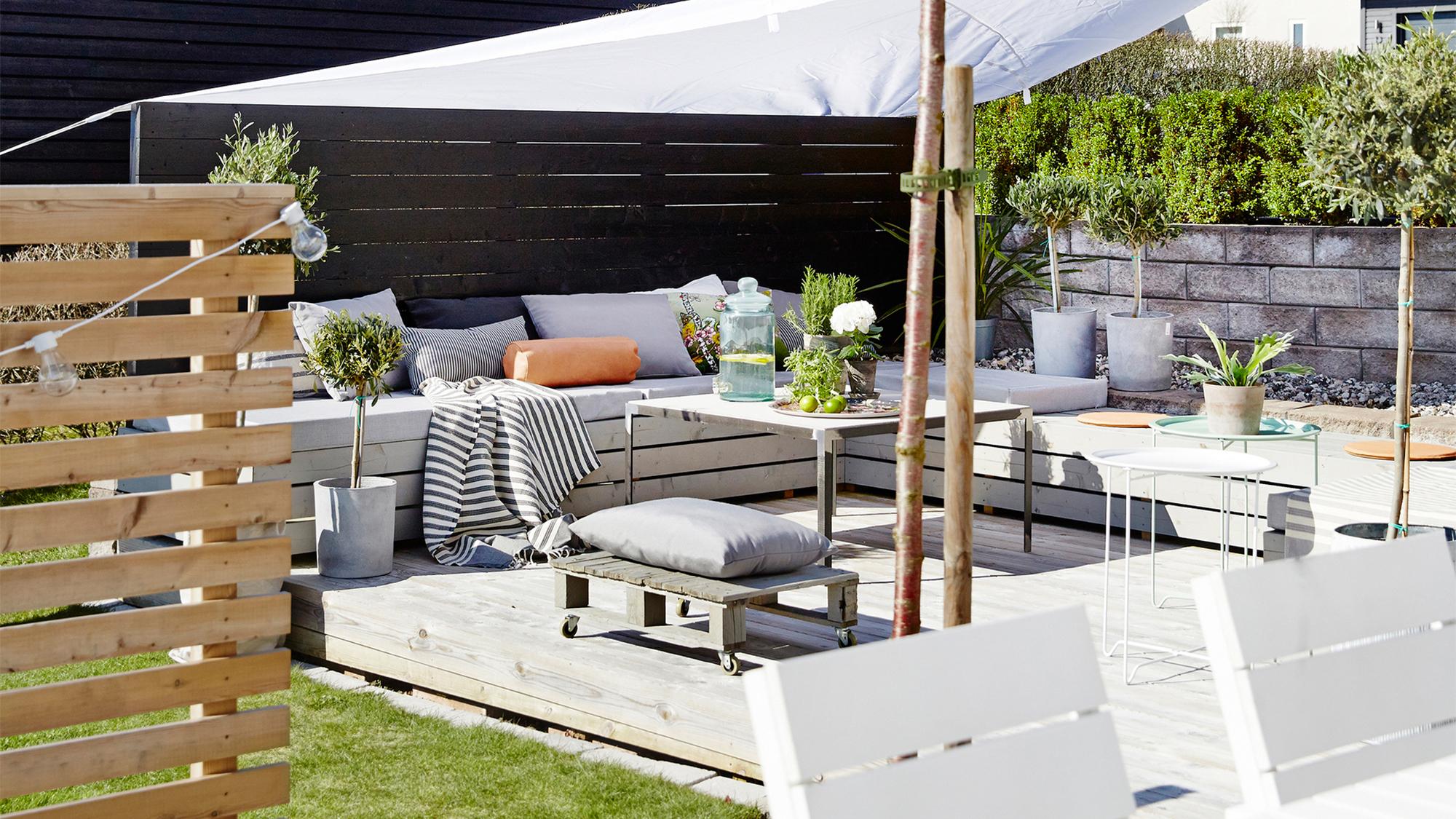 An outdoor light wood deck with built-in cushioned seating, a table with drinks and potted plants, under a white shade sail, next to a slatted wooden fence.