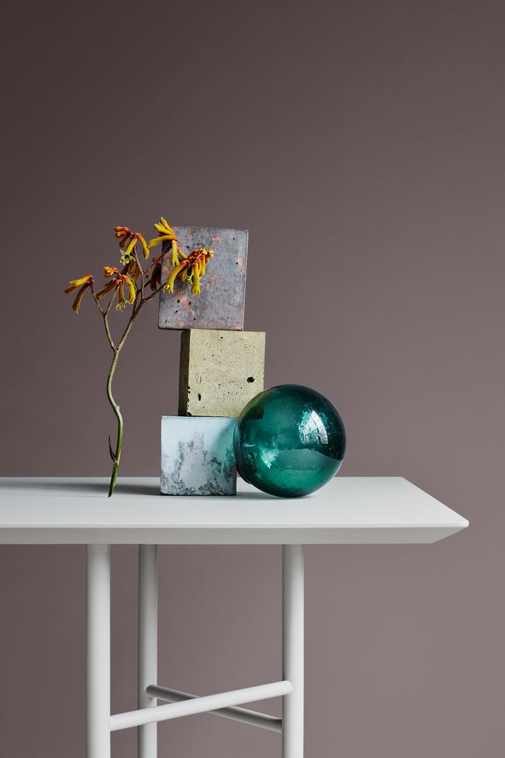 A still life with a branch of yellow-orange flowers, a stack of three textured blocks (dark metal, concrete, marbled), and a large green glass orb on a white table against a brown wall.