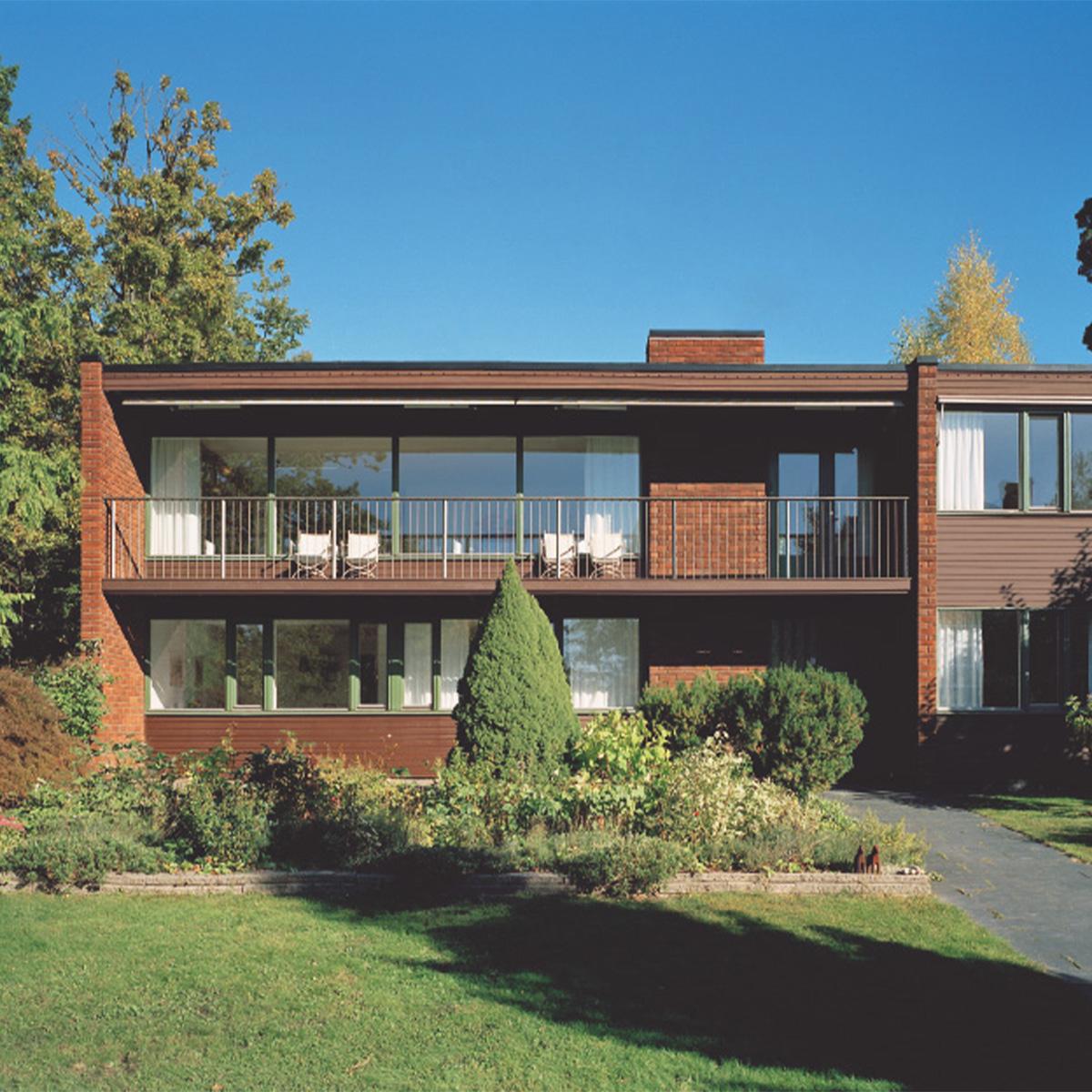 Two-story dark brown building with brick accents, a balcony with white chairs, a garden, and trees under a blue sky.