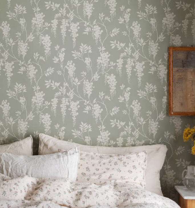 Sage green wallpaper with a white wisteria pattern above a bed with neutral and floral bedding.