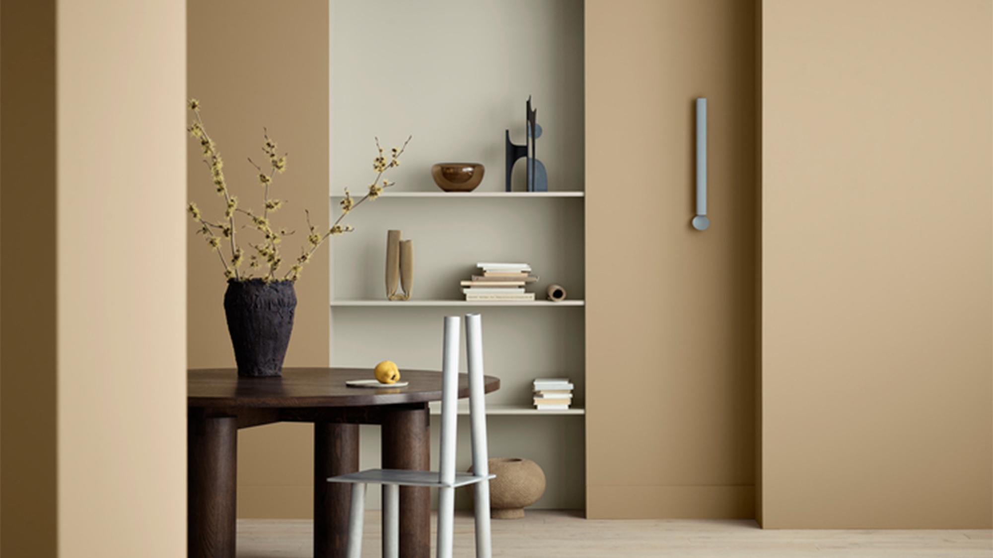 A modern room with tan walls, a dark round table holding a vase of yellow branches, a light grey chair, a built-in shelf unit, and a pale blue handle on the wall.