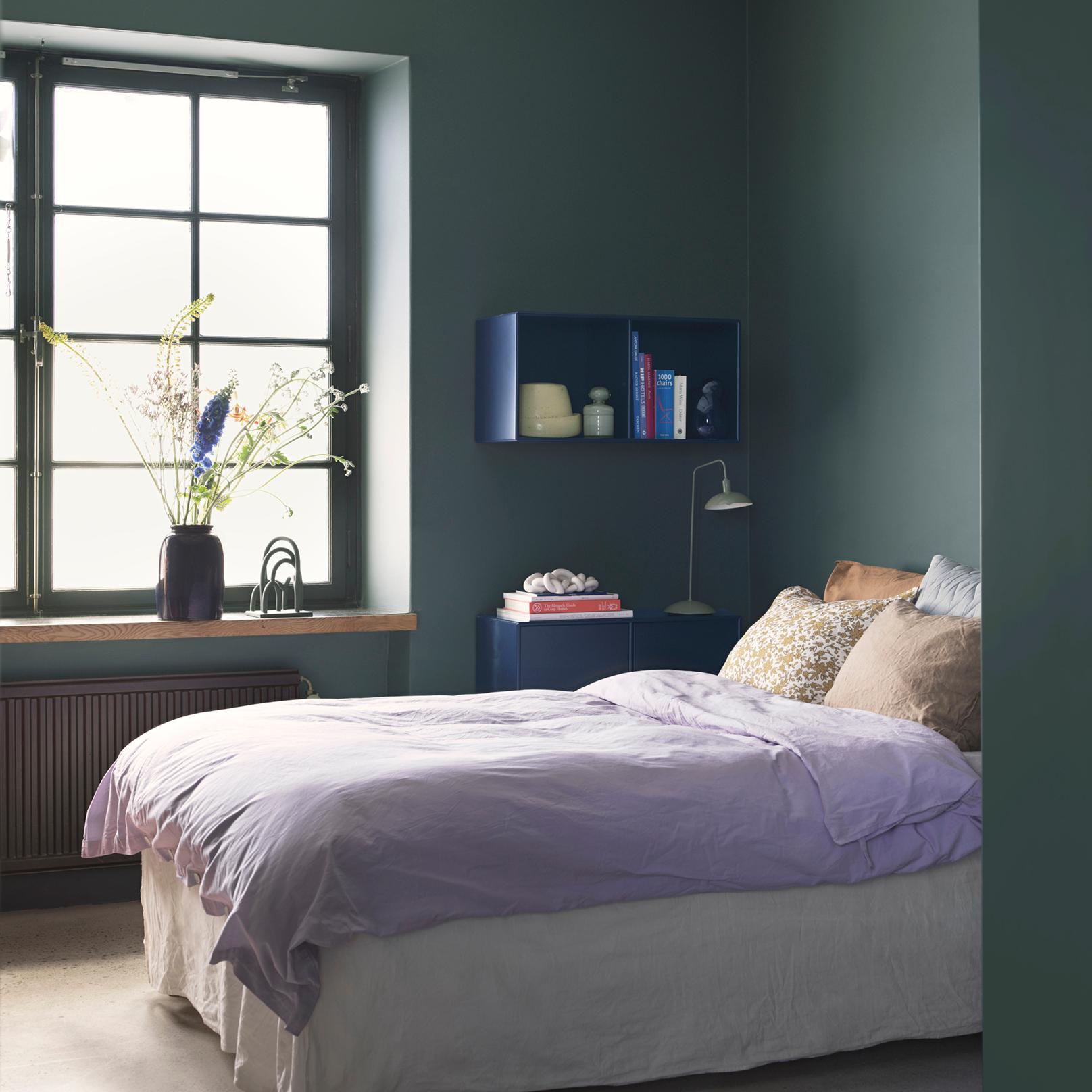 A bedroom with dark green walls, a lavender duvet on the bed, and a window displaying flowers.