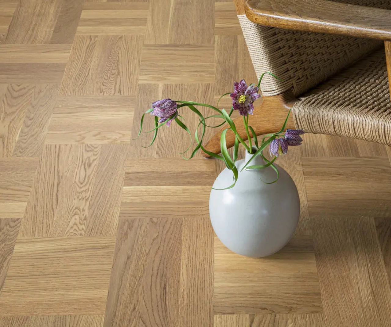 White vase with purple flowers on a light parquet wood floor next to a woven chair.