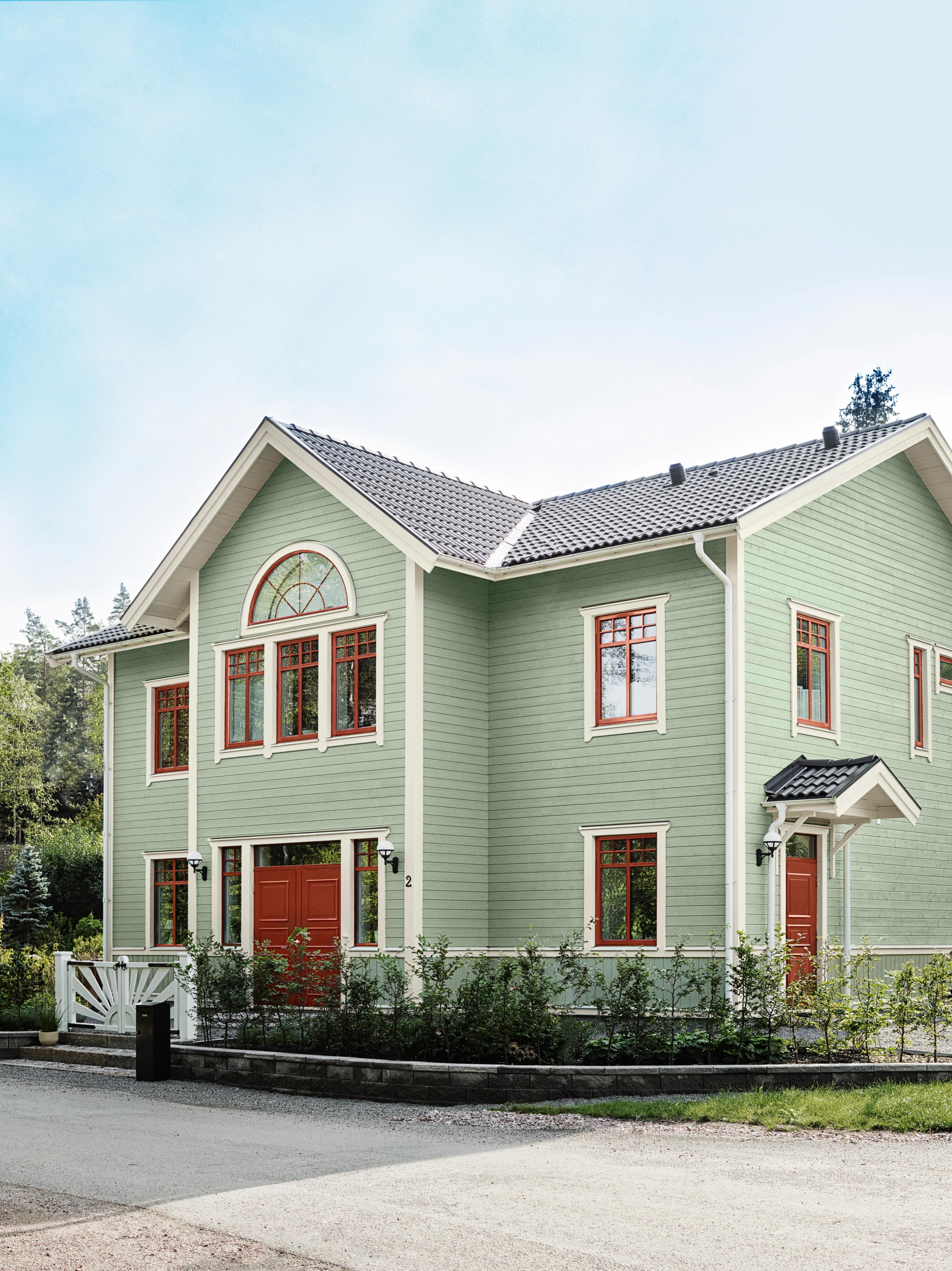 A large, two-story house with pale green siding, red-framed windows and doors, and a dark pitched roof, seen under a clear blue sky.