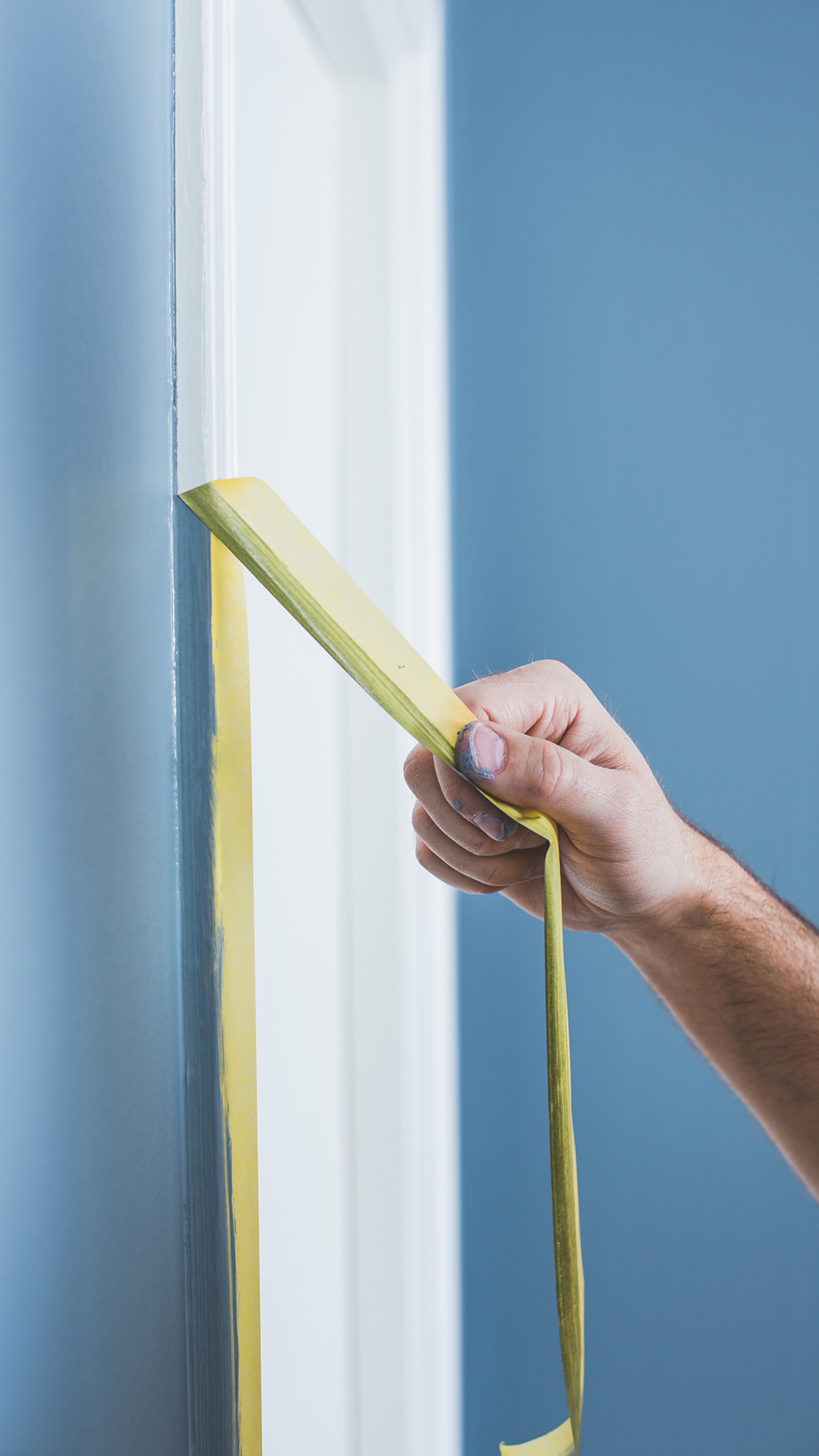 Hand peeling yellow painter's tape from a blue wall next to white trim.